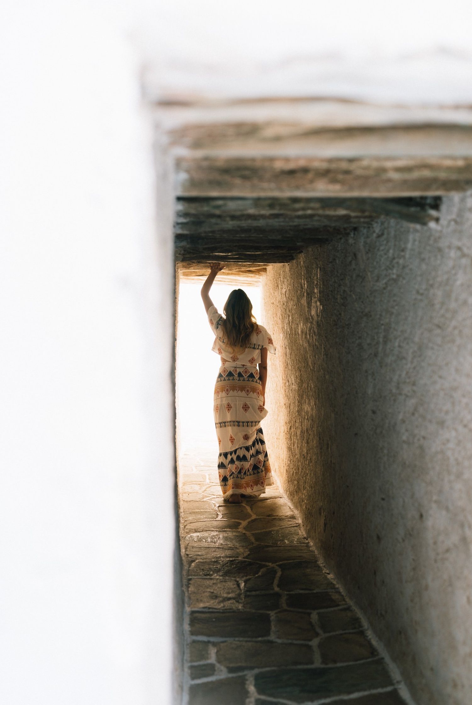 A silhouetted figure walking through a narrow stone passageway towards bright sunlight.