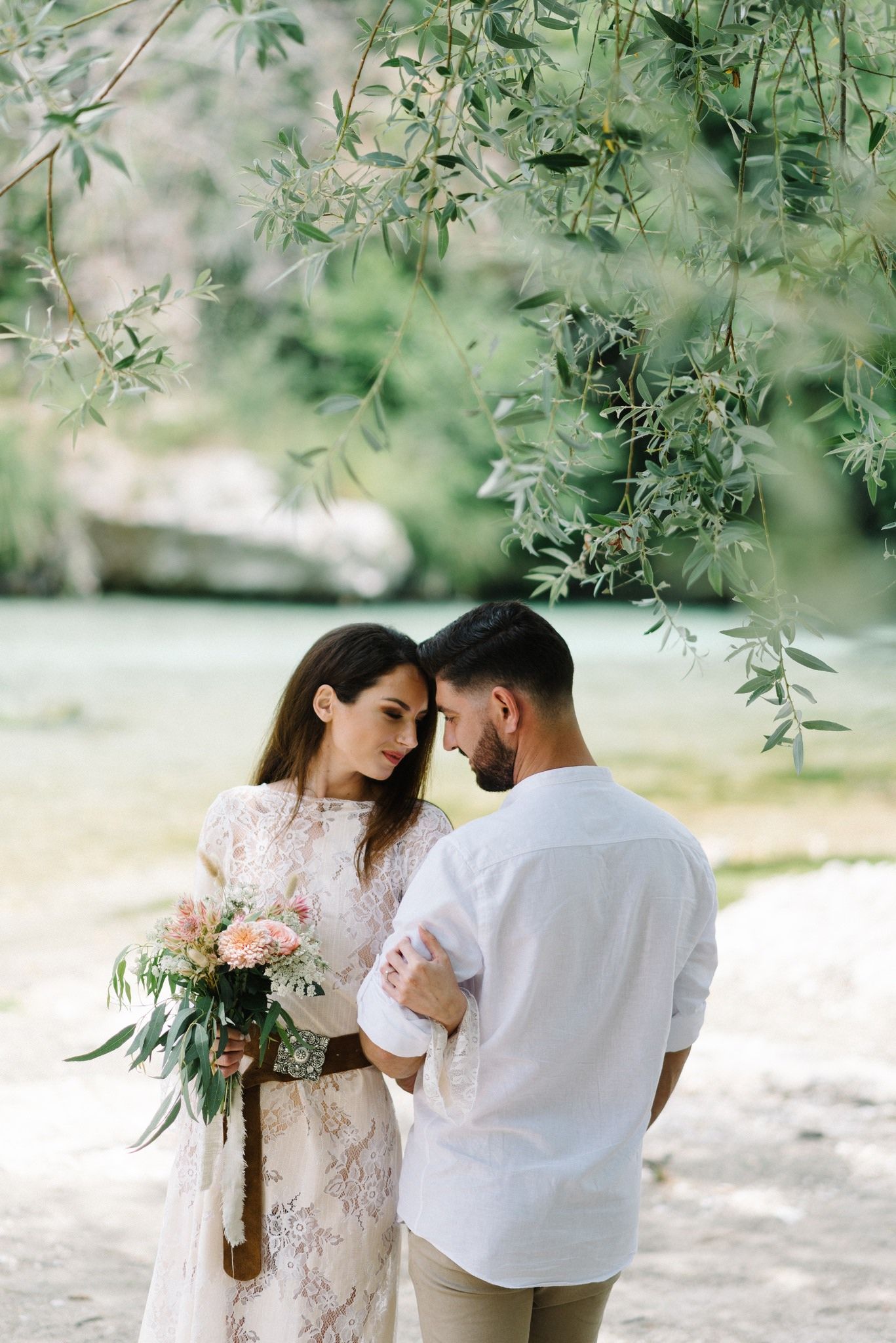 Romantic couple embracing under hanging tree branches with delicate bouquet.