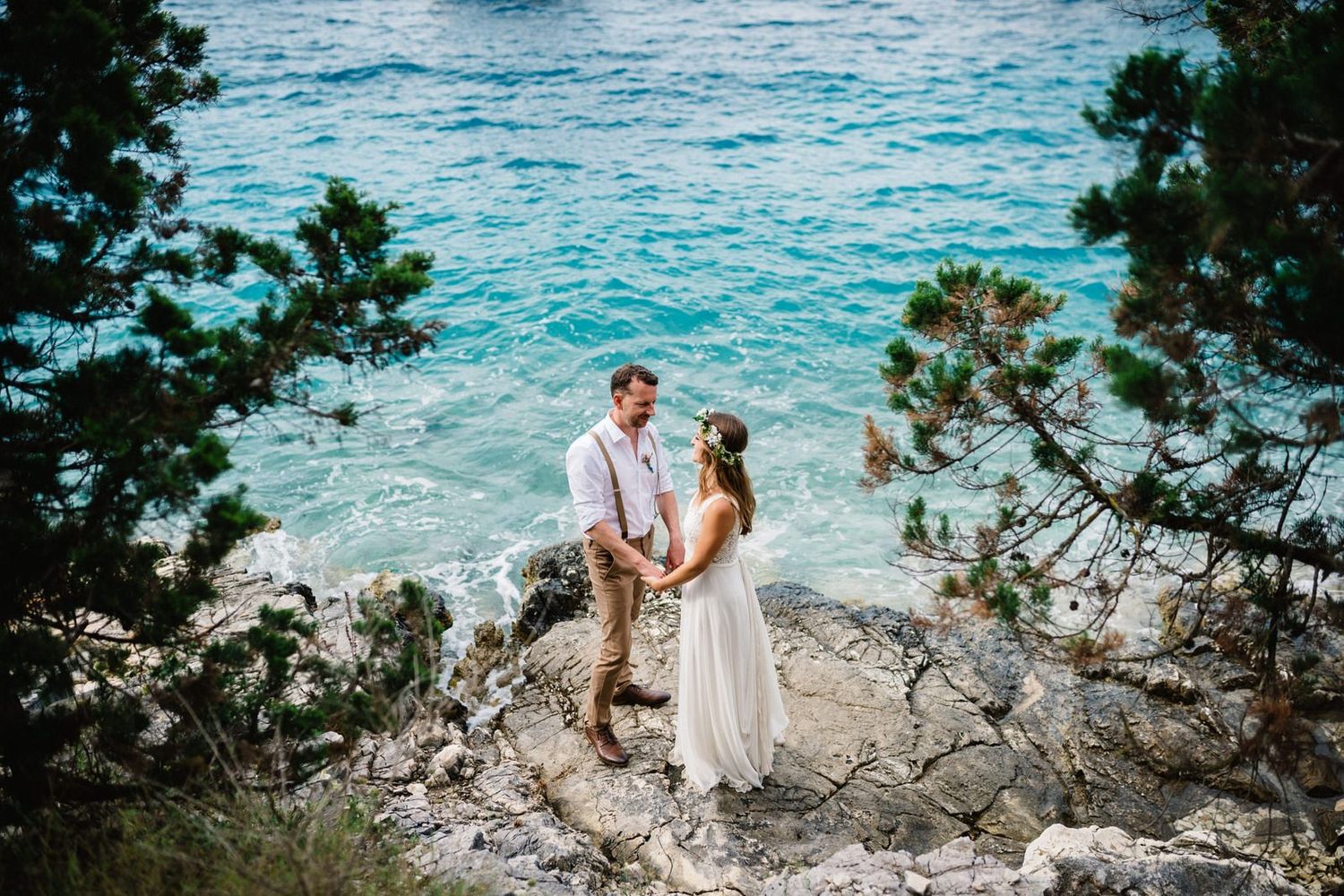 Newlyweds standing on rocky coastal cliffs overlooking turquoise Mediterranean waters.