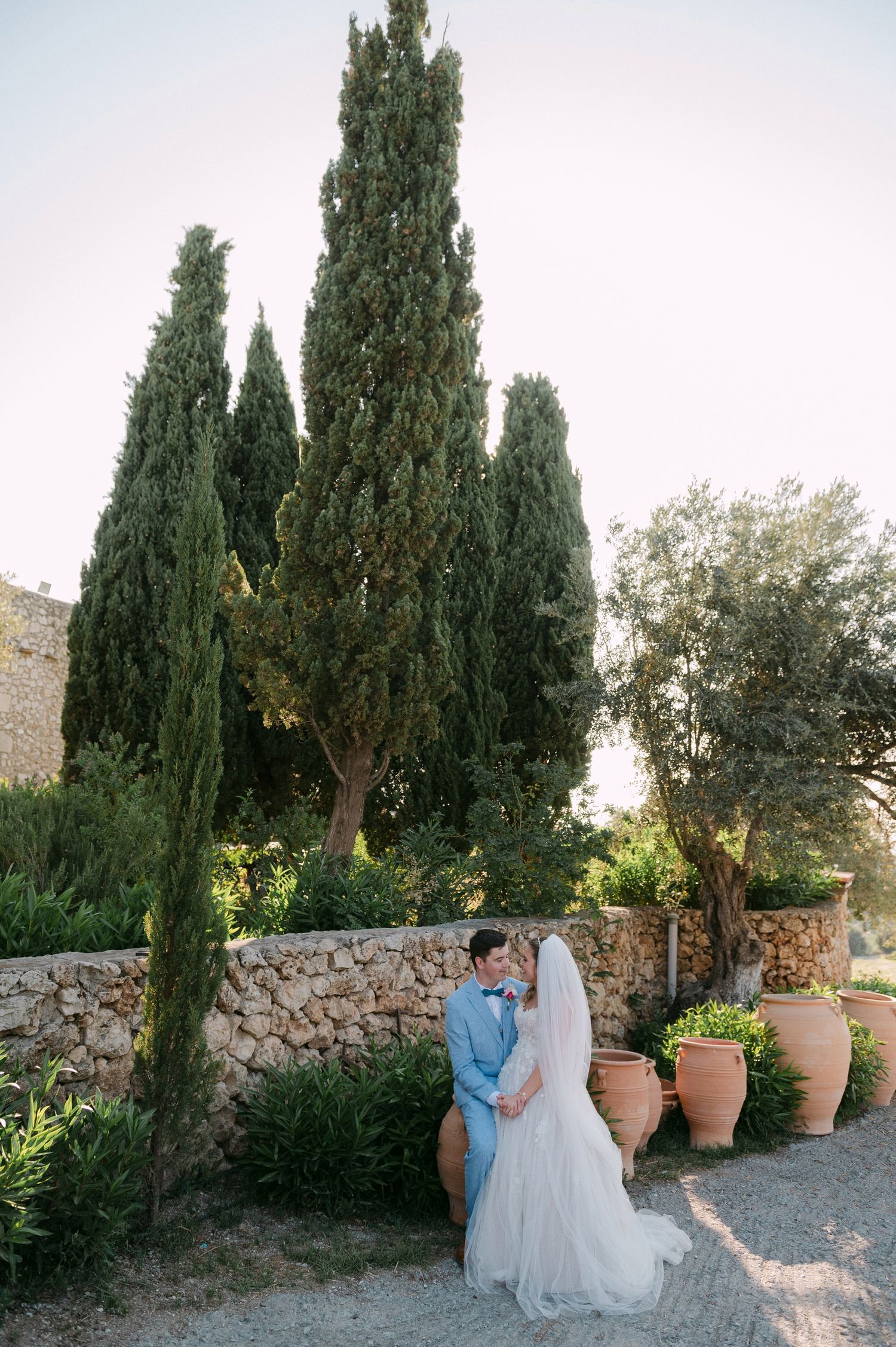A wedding couple stands near tall cypress trees and stone walls with terracotta pots in a Mediterranean garden setting.