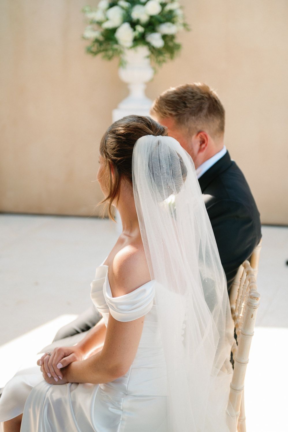 A bride in a white off-shoulder dress and long veil sits beside someone in a black suit during an intimate wedding moment.