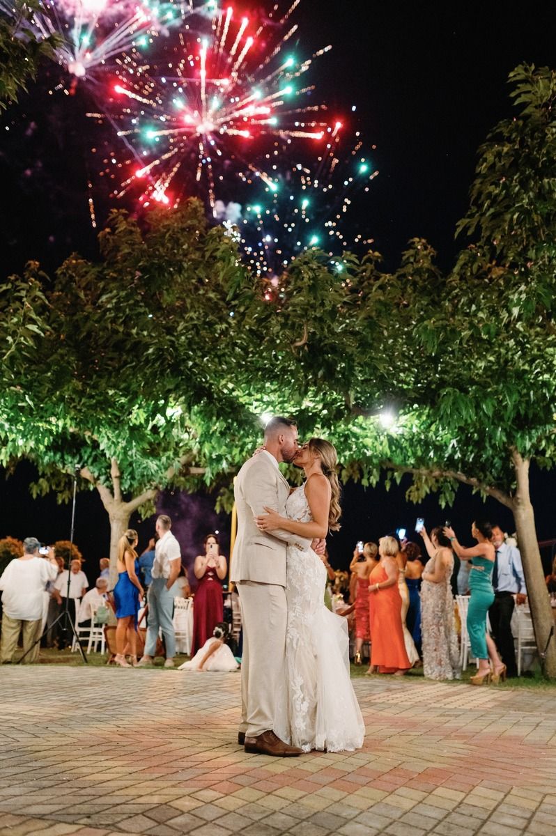 Colorful fireworks burst over a nighttime wedding celebration as a couple dances under trees.