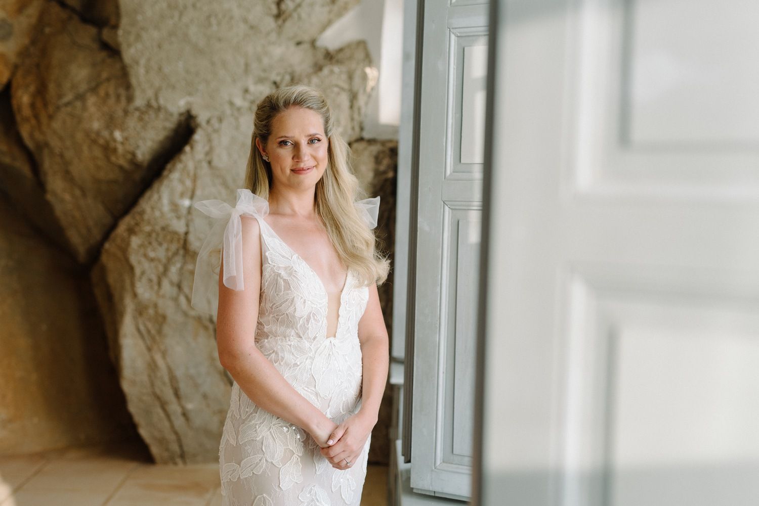 A person in an intricate white lace wedding gown stands near a window with natural lighting.