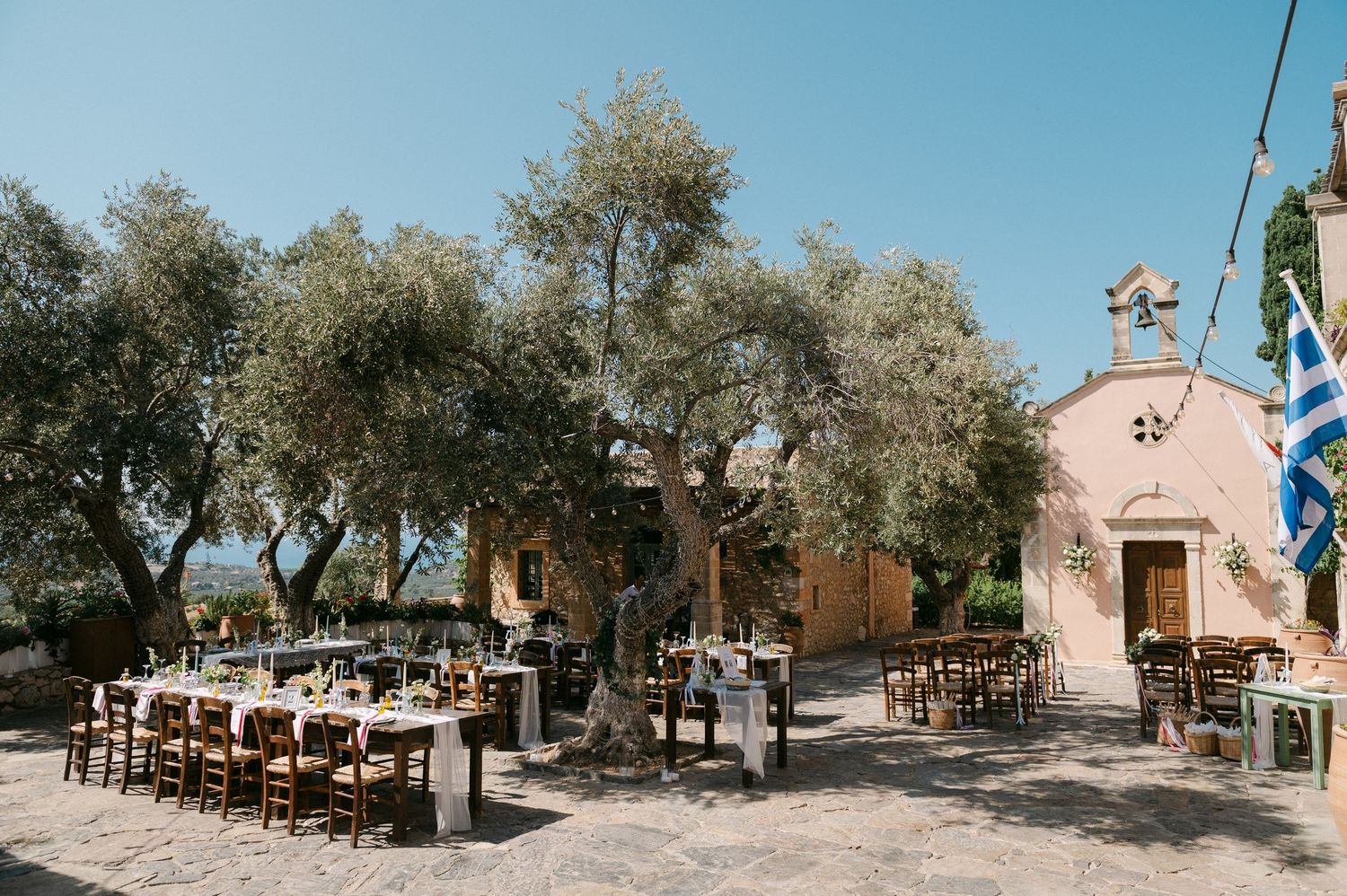 Rustic outdoor wedding ceremony setup with wooden chairs arranged beneath olive trees in a Mediterranean setting.