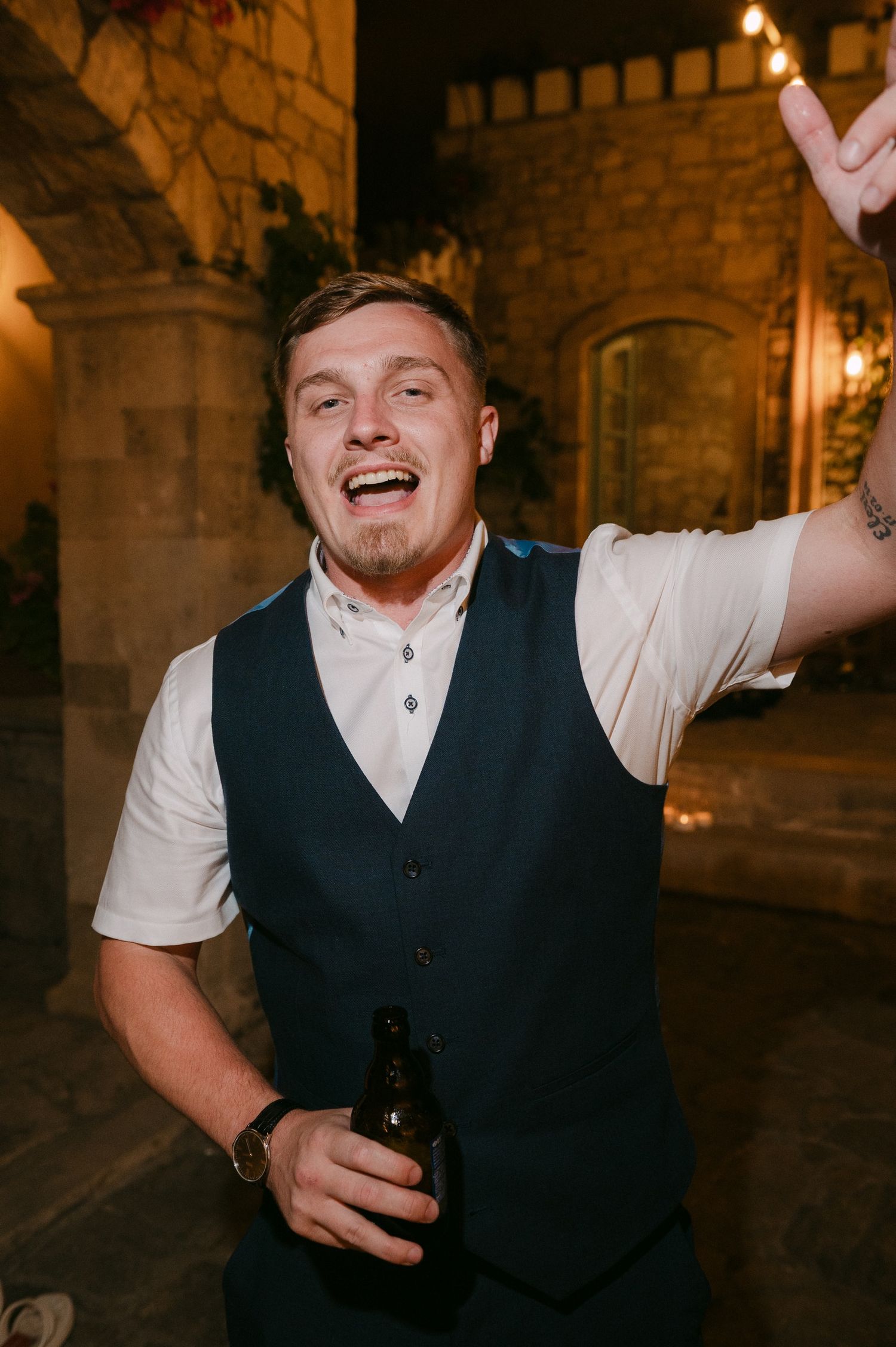 A joyful person in a dark vest holds a bottle during an evening celebration at a stone-walled venue.