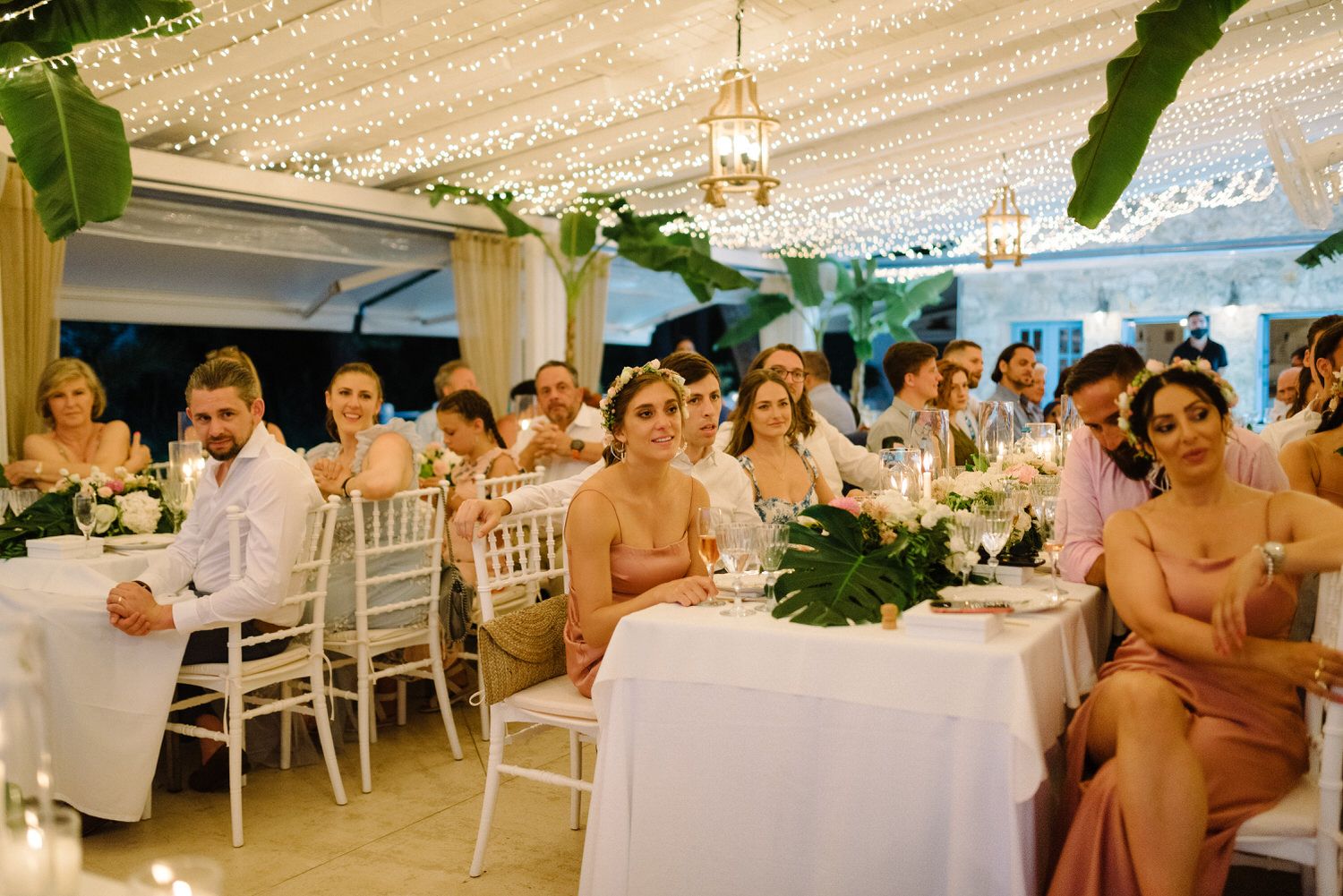 Wedding guests seated at white tables under a draped tent with floral centerpieces.