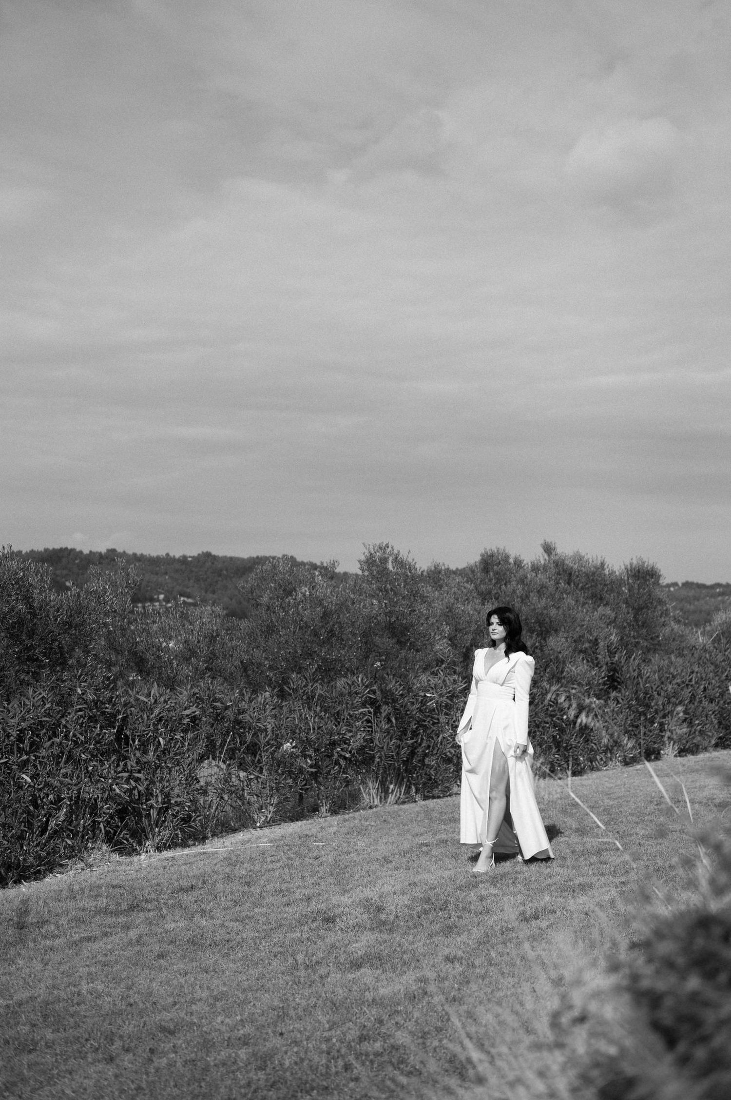 Two figures in white dresses stand together on a grassy hillside under cloudy skies in black and white photography.