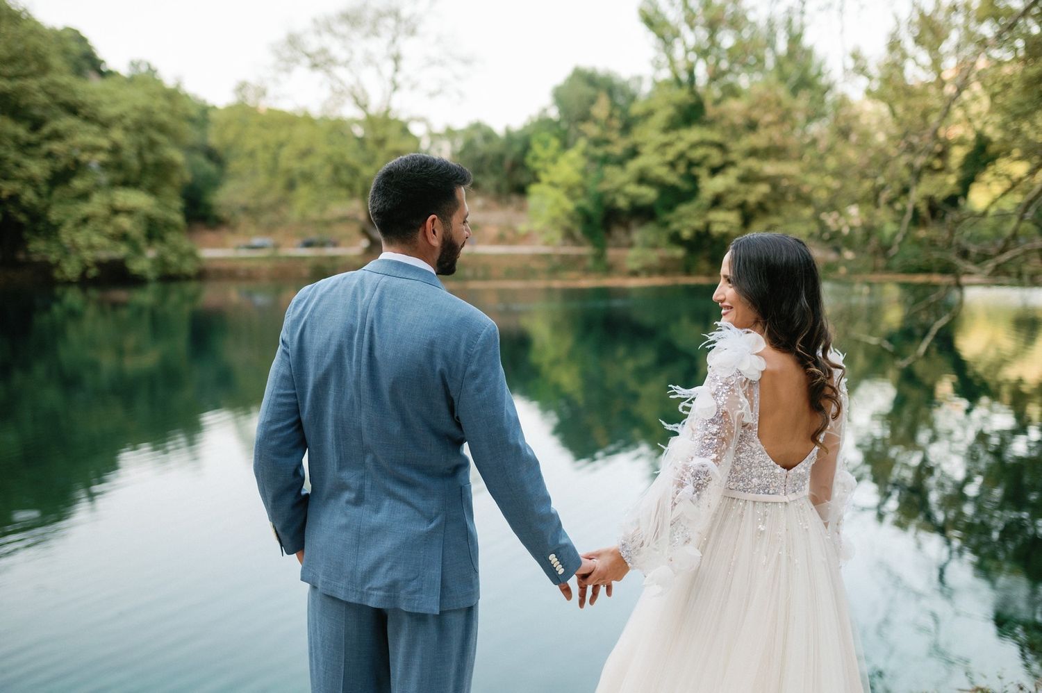 A couple in wedding attire holds hands while standing at the edge of a serene lake surrounded by green trees.