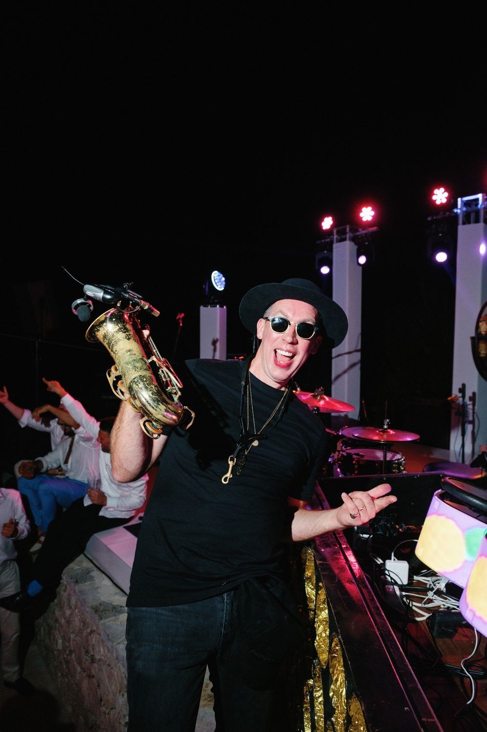 Performer in black attire and sunglasses holds up a gold saxophone while gesturing enthusiastically at a nighttime event.