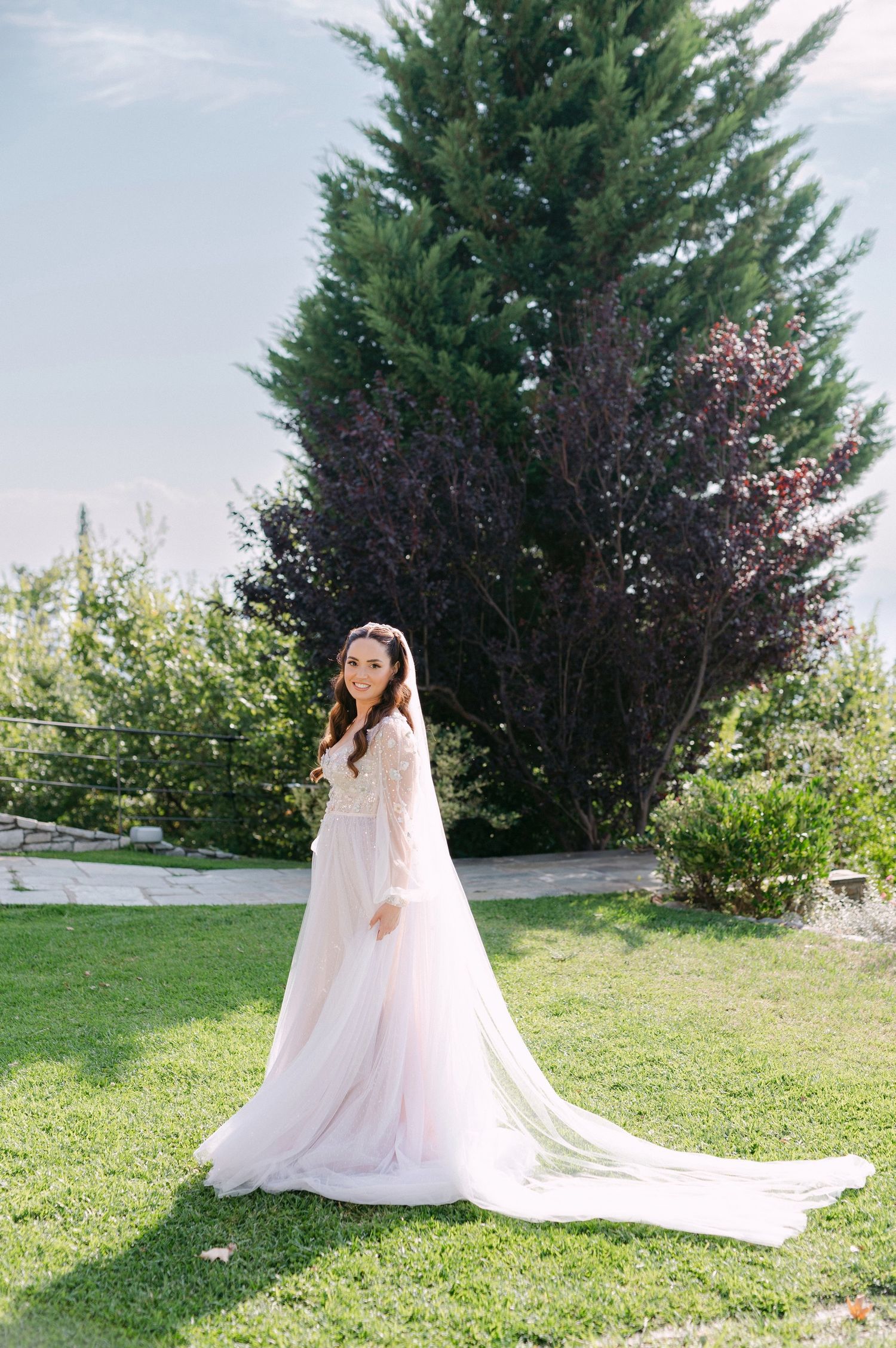 A flowing white wedding dress with train sweeps across a manicured lawn with evergreen trees in the background.
