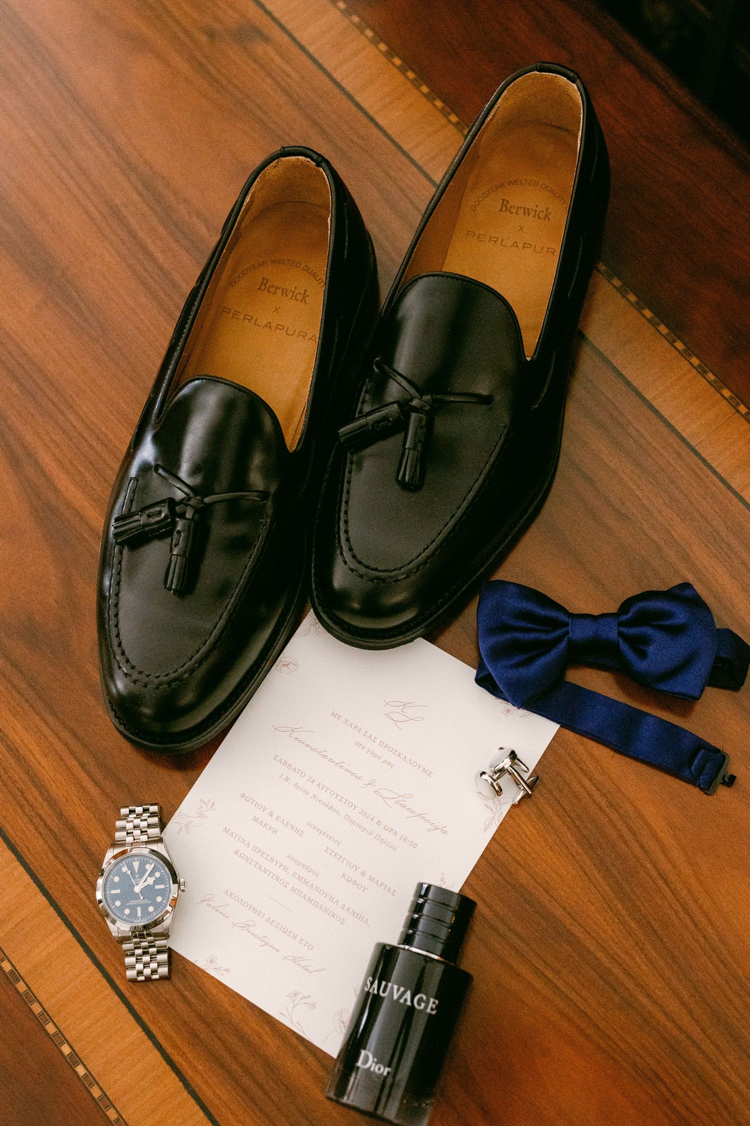 Black leather tassel loafers displayed with a blue bowtie, wristwatch, cologne bottle and paper on wooden flooring.