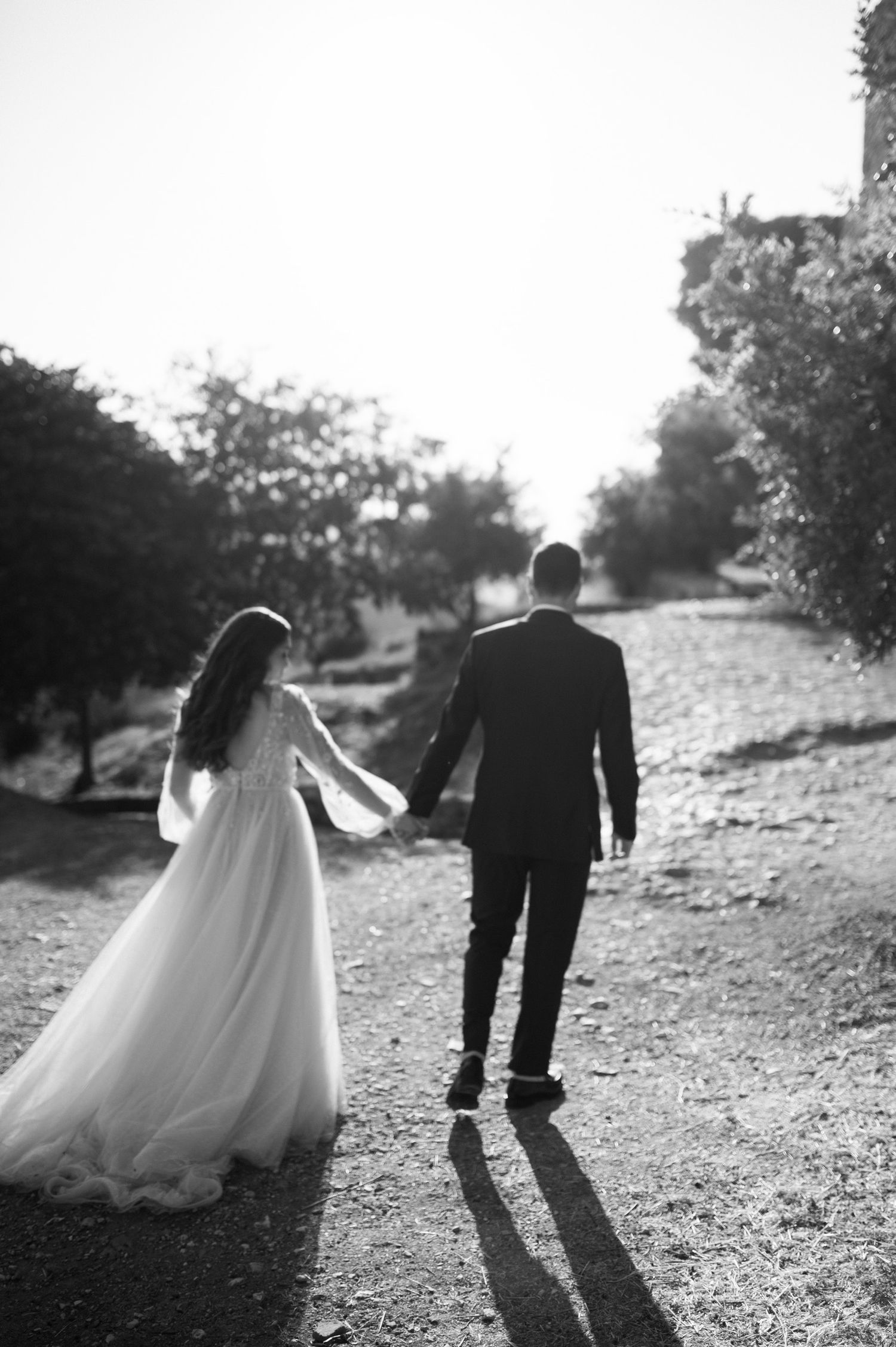 A black and white photo of a couple in wedding attire walking together on a sunlit dirt path.