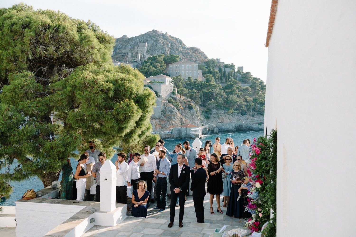 Wedding guests gather on scenic coastal terrace with mountain and ocean views in background.