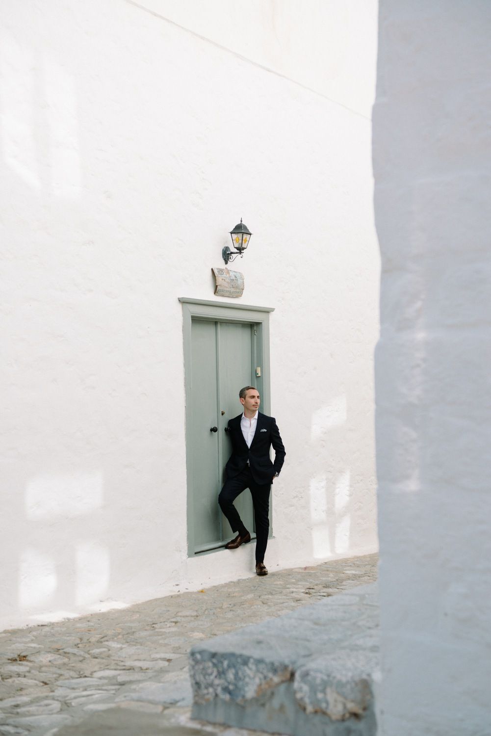 A person in formal attire poses against a white wall with a mint green door.