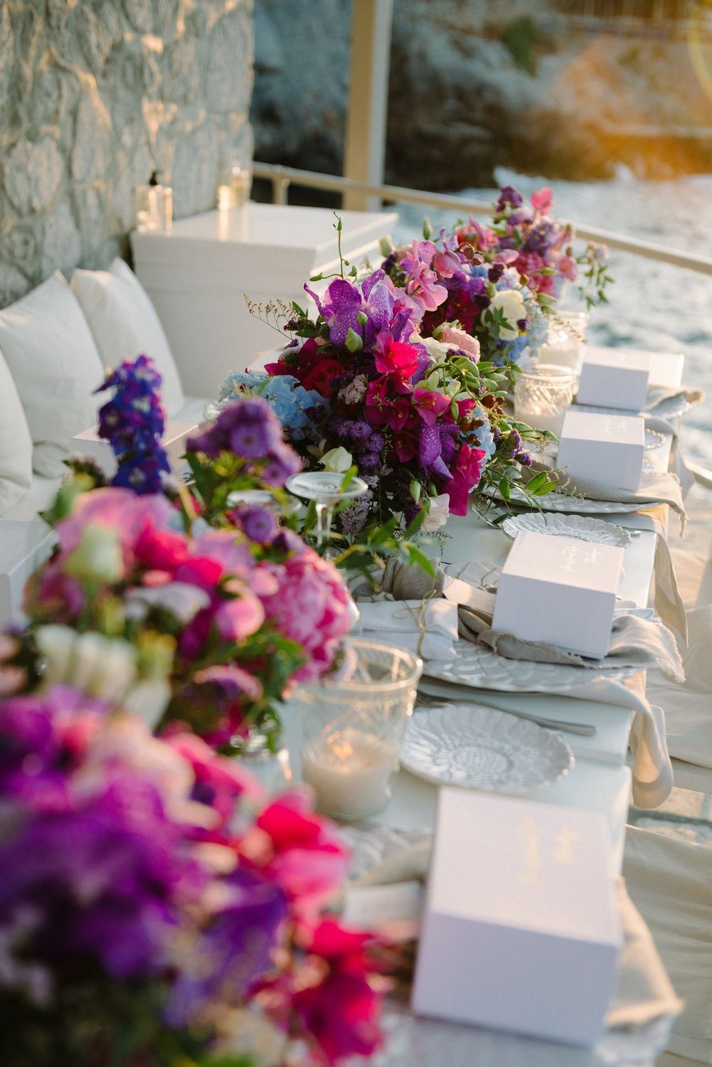 Coastal dining setup with pink and purple floral arrangements and white place settings at sunset.