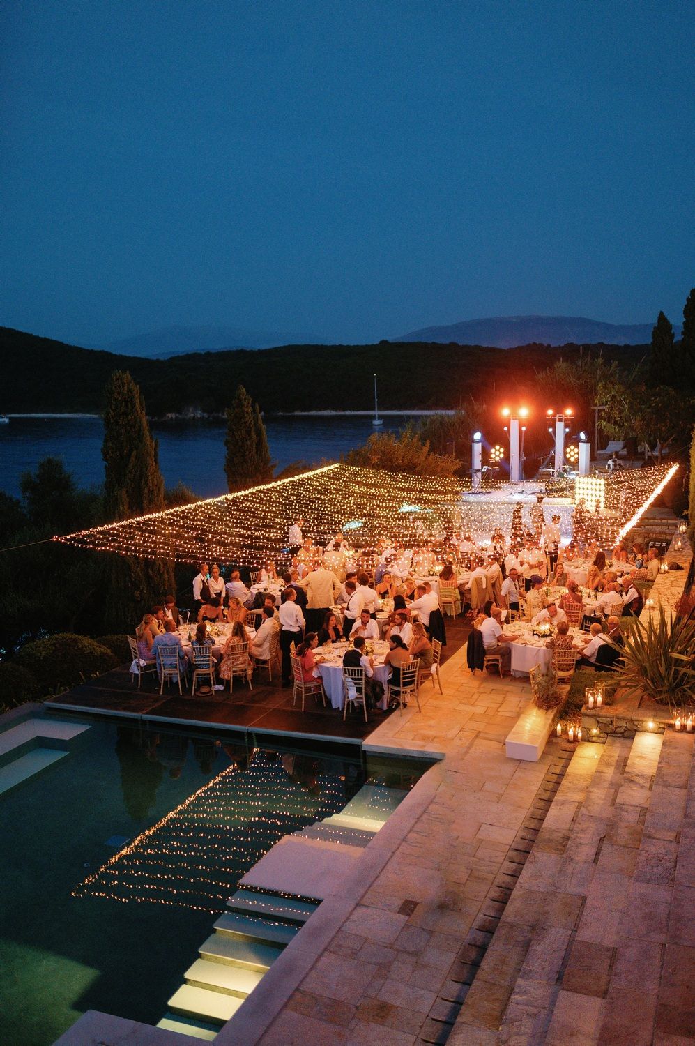 Outdoor wedding reception at dusk with strings of lights illuminating guests dining by a pool overlooking water.