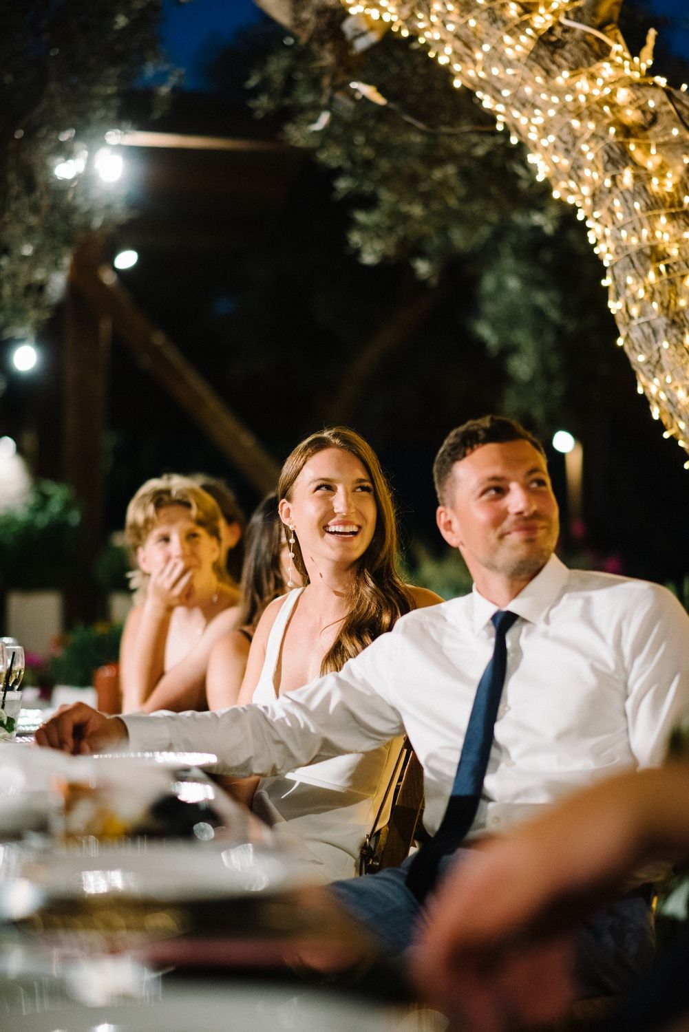 Wedding guests enjoy a candlelit outdoor reception dinner under twinkling lights and trees.