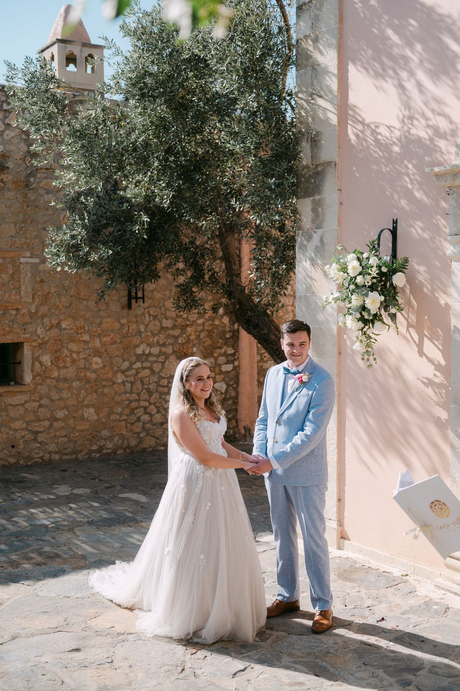 Romantic Mediterranean wedding couple shares intimate moments against pink stucco walls and olive trees in sunlit courtyard.