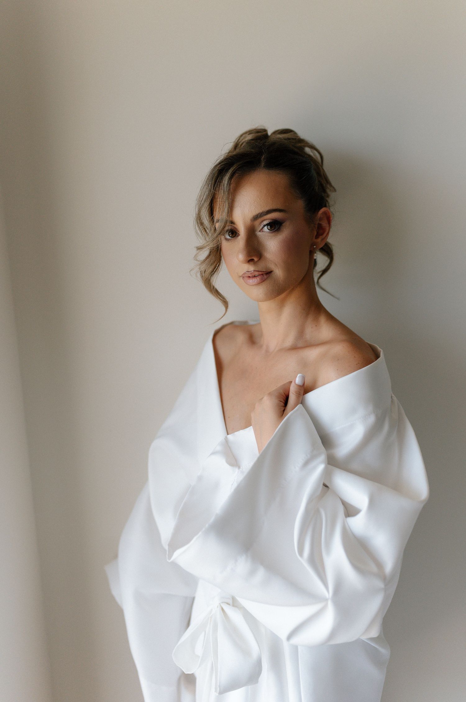 A bride in a white satin robe with an off-shoulder look, elegant updo, and natural makeup poses gracefully.