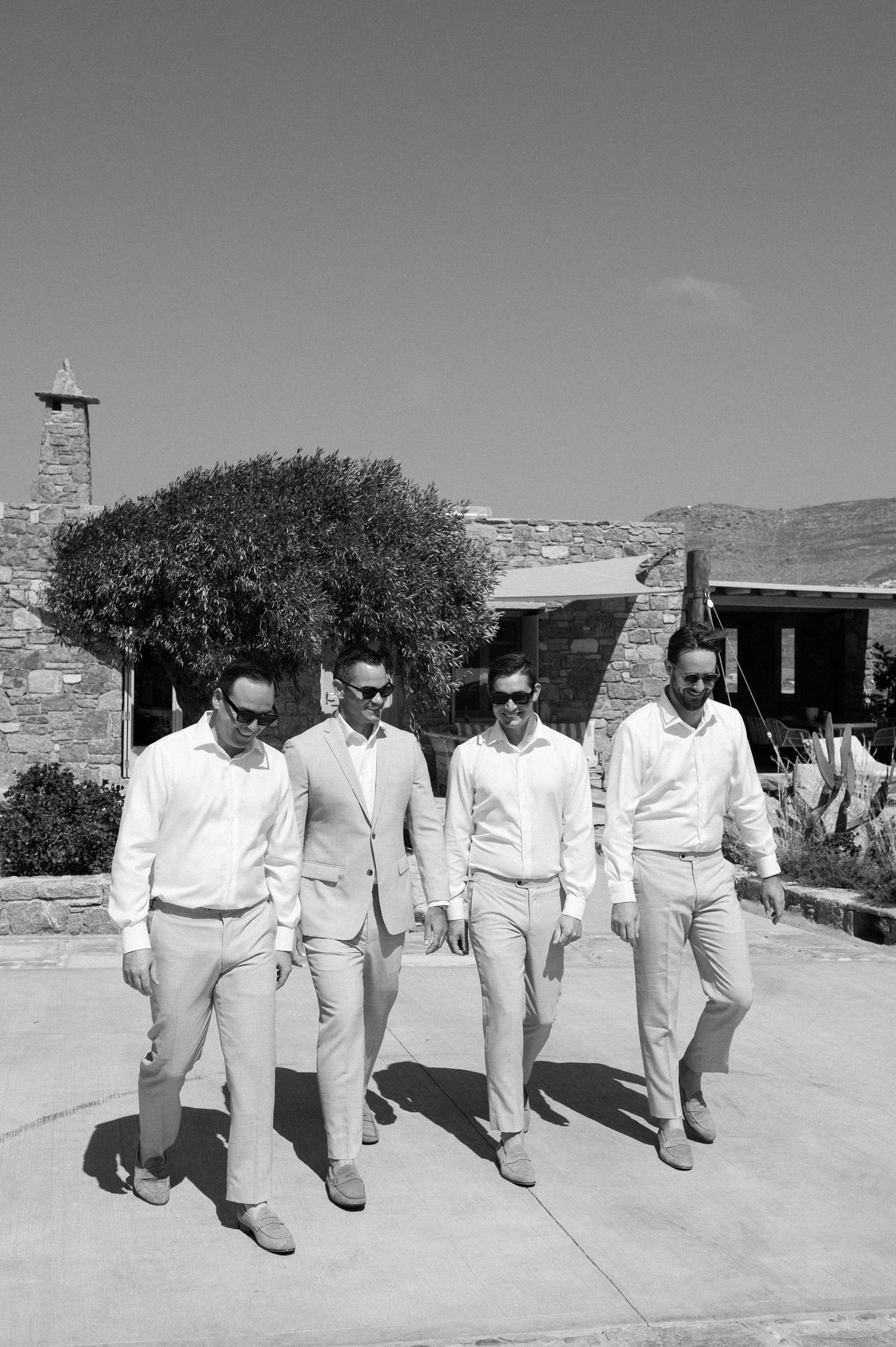 Four men in light-colored clothing walk together in a line on a patio with stone walls and shrubs in black and white.