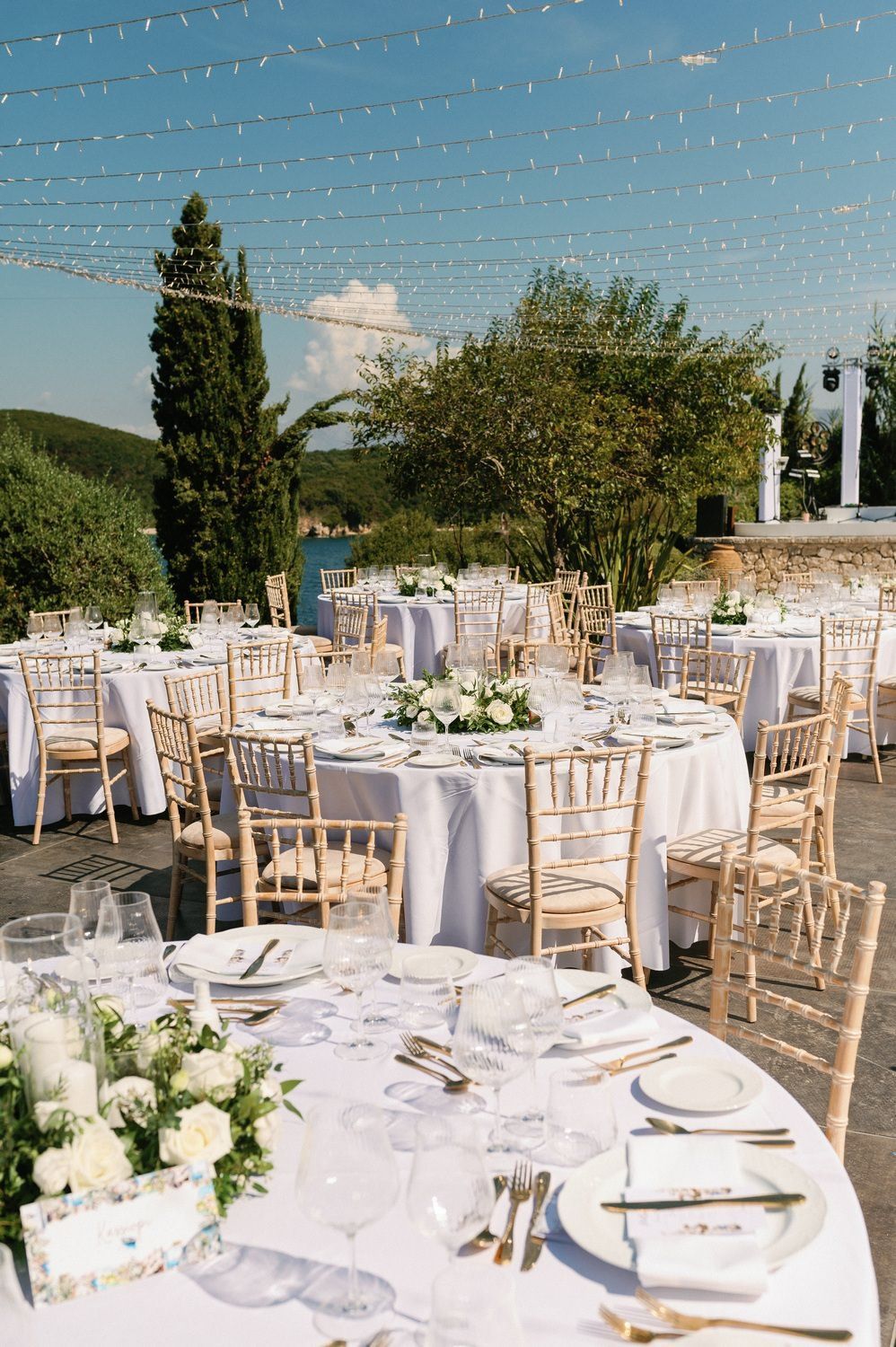 Elegant outdoor wedding reception setup with white-clothed tables, chiavari chairs, and string lights overhead.