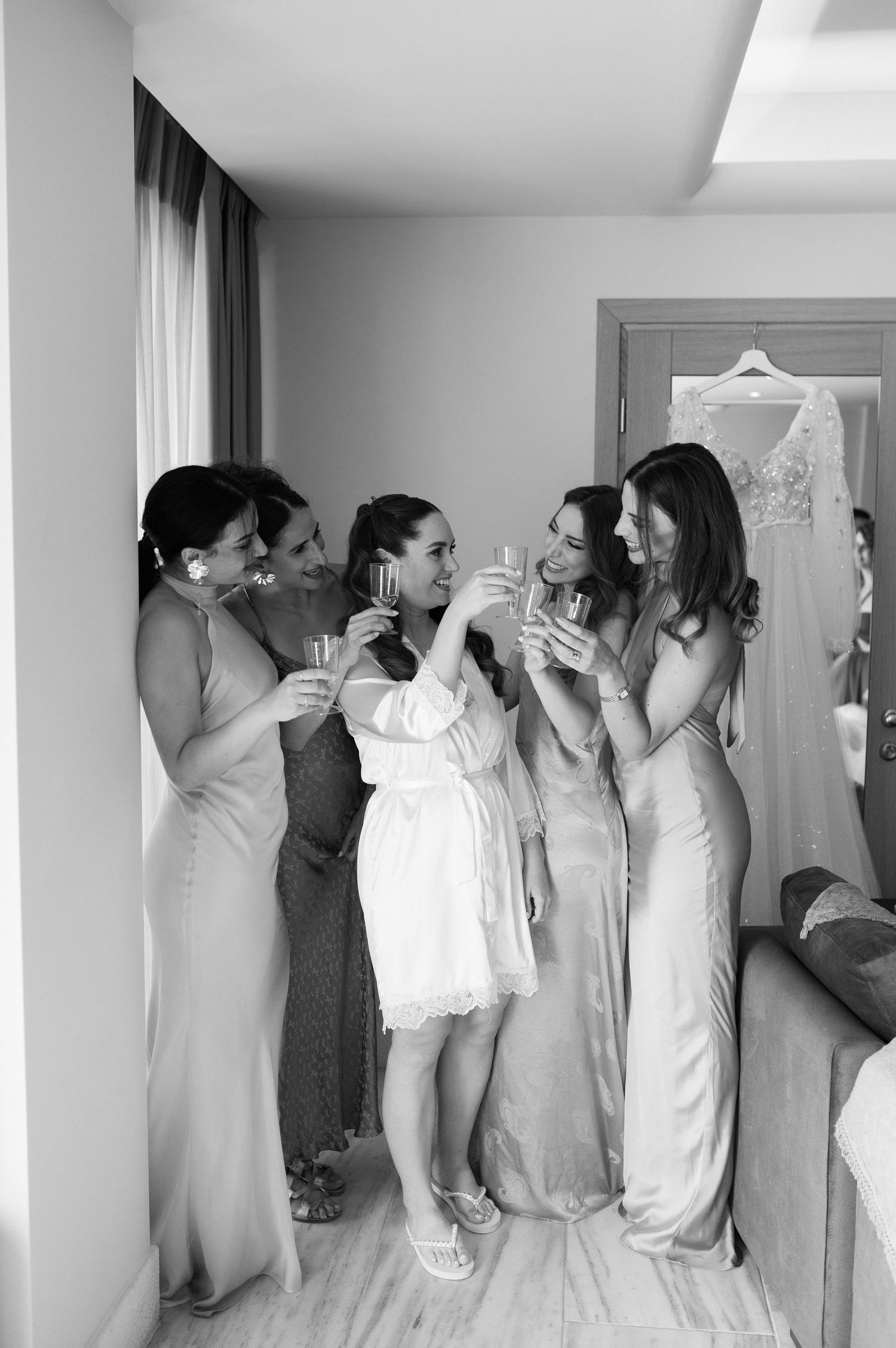 A group in formal attire toasts with champagne glasses in a bridal suite with a white dress visible hanging in background.