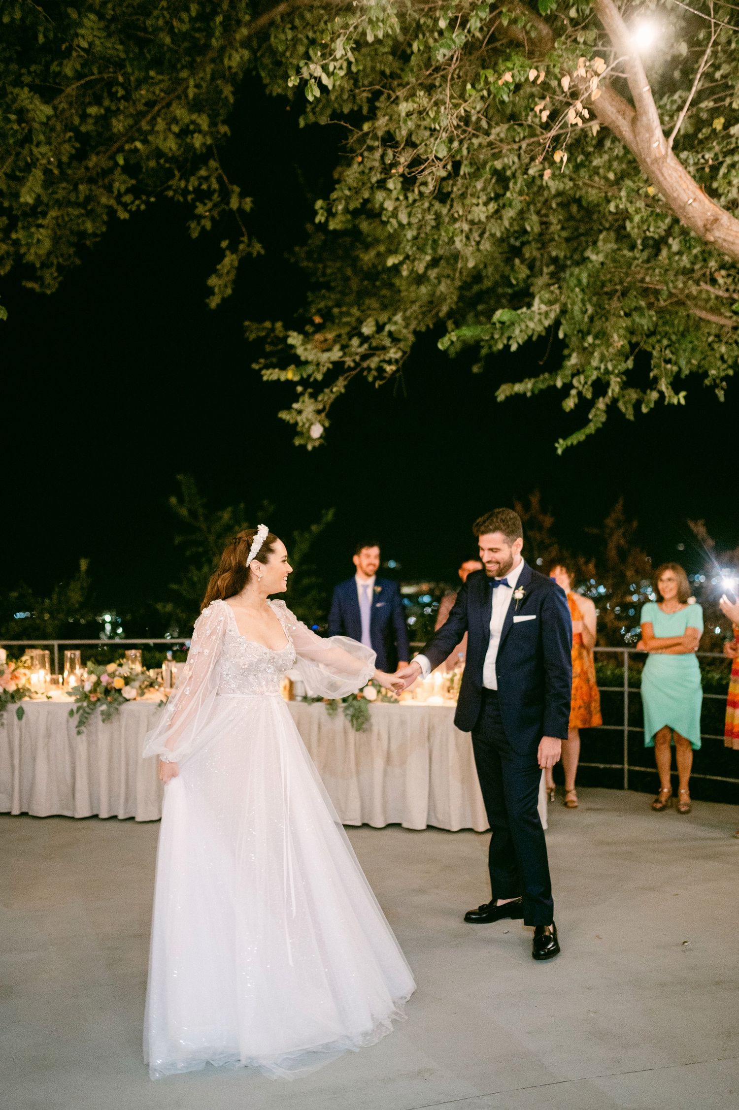 A couple shares their first dance at an outdoor evening wedding reception under trees with string lights.