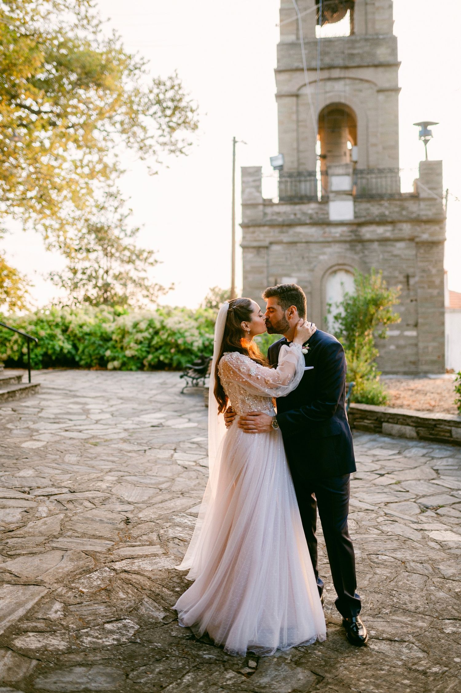 A couple shares a romantic moment in front of a stone bell tower during golden hour on their wedding day.