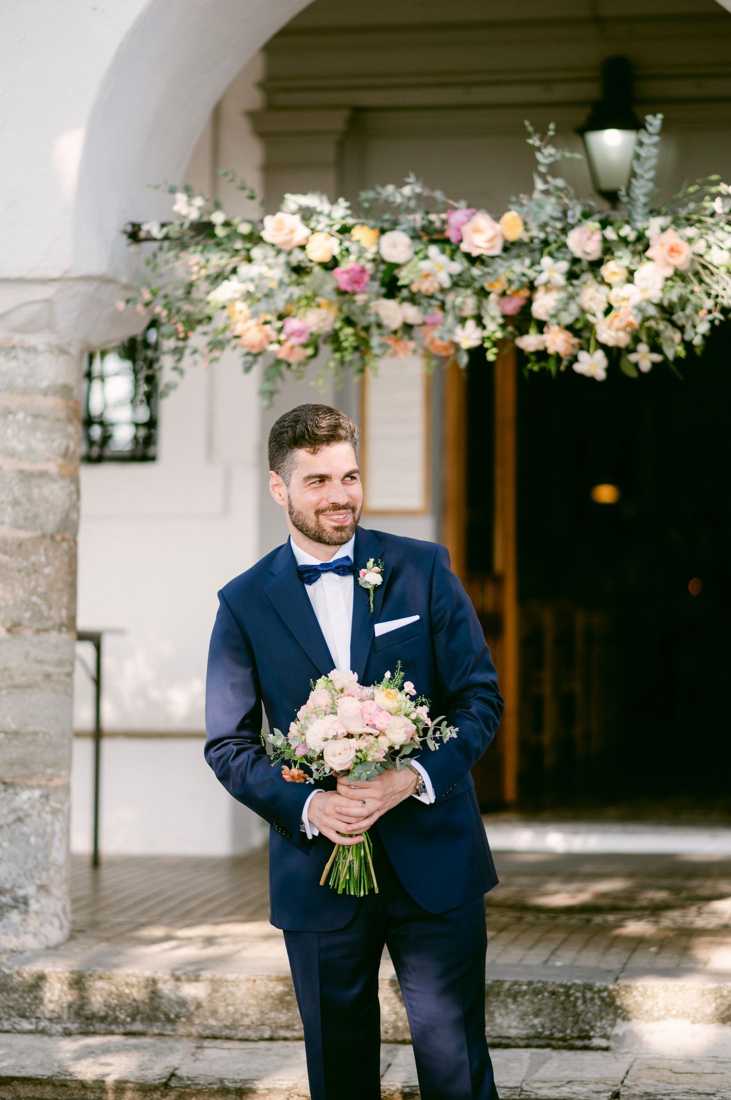 Elegant wedding attire with navy suit and bow tie complemented by soft pink and white floral bouquet under arched entrance.