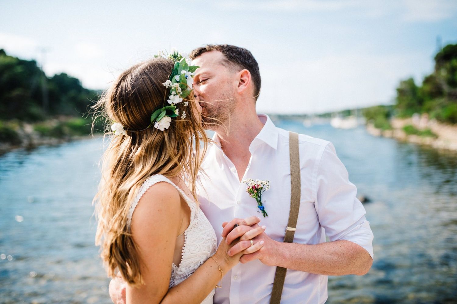 A romantic wedding couple embracing by a scenic waterfront with boats in the background.