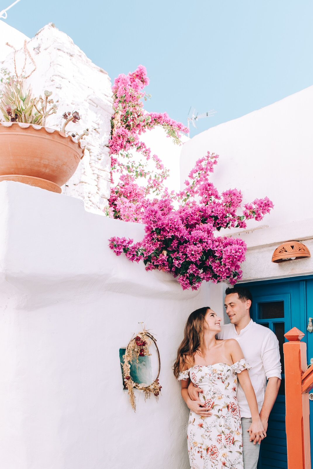 Romantic couple poses under pink bougainvillea by turquoise door in Mediterranean setting.