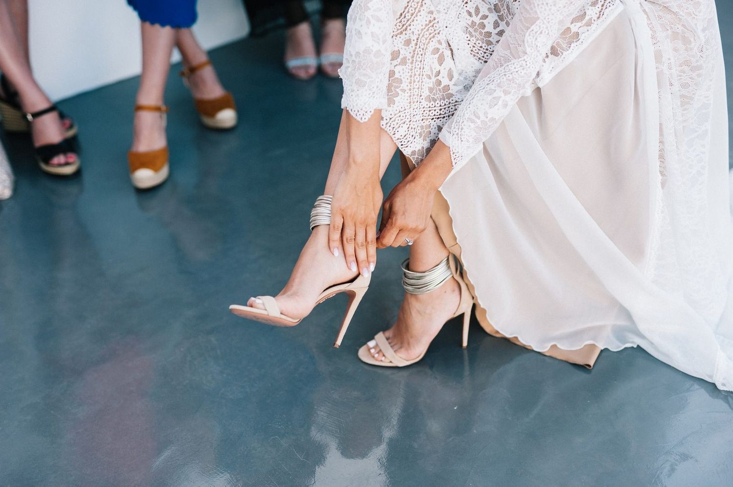 Close-up of bride putting on elegant white heeled sandals on concrete floor.