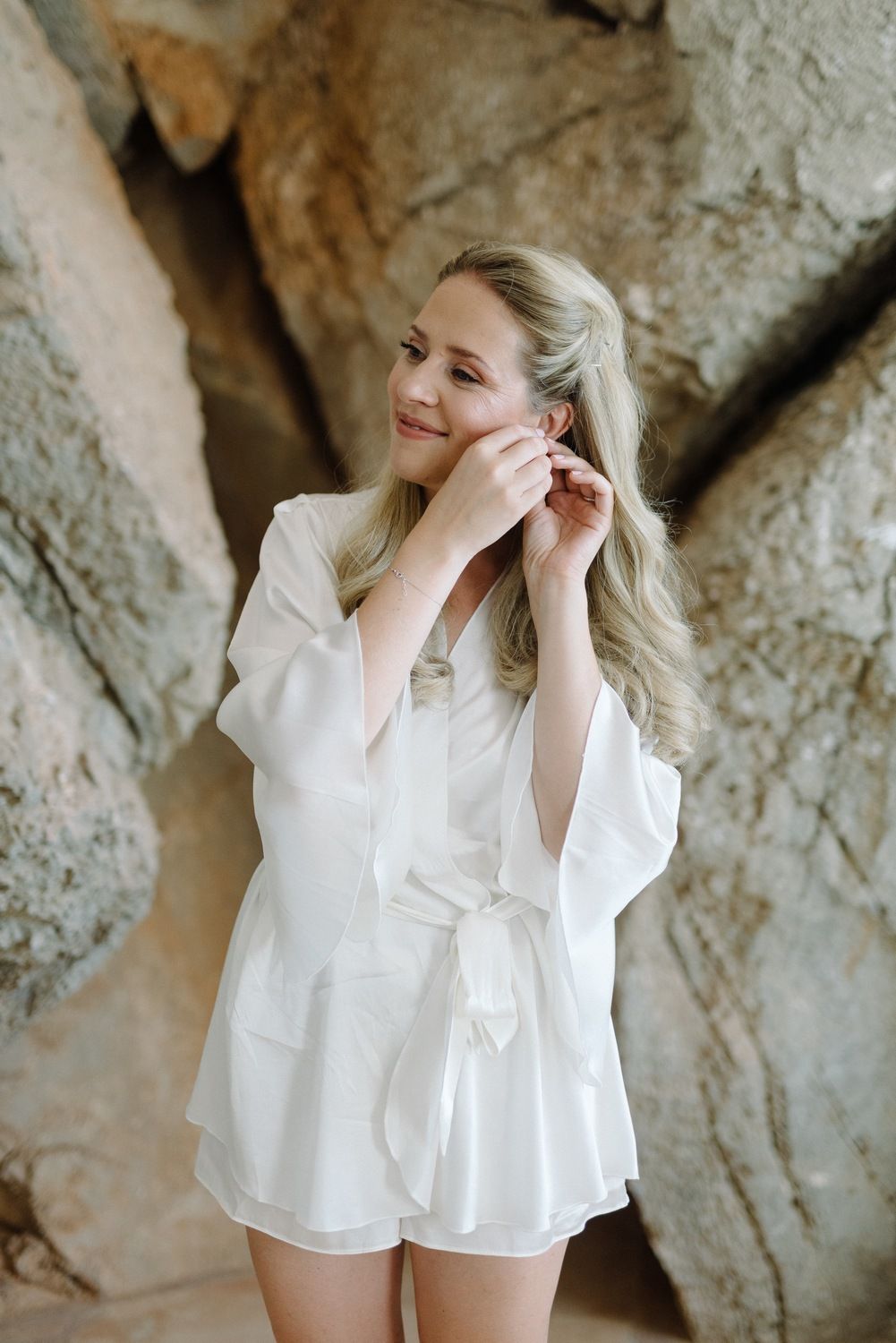 A woman in a white summer romper poses gracefully against a textured rock wall outdoors.