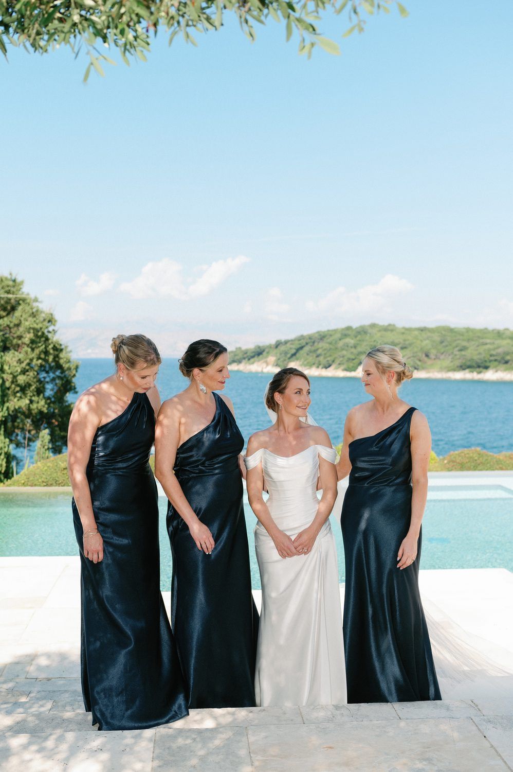 A bride in white stands with bridesmaids in navy dresses against a coastal Mediterranean backdrop.