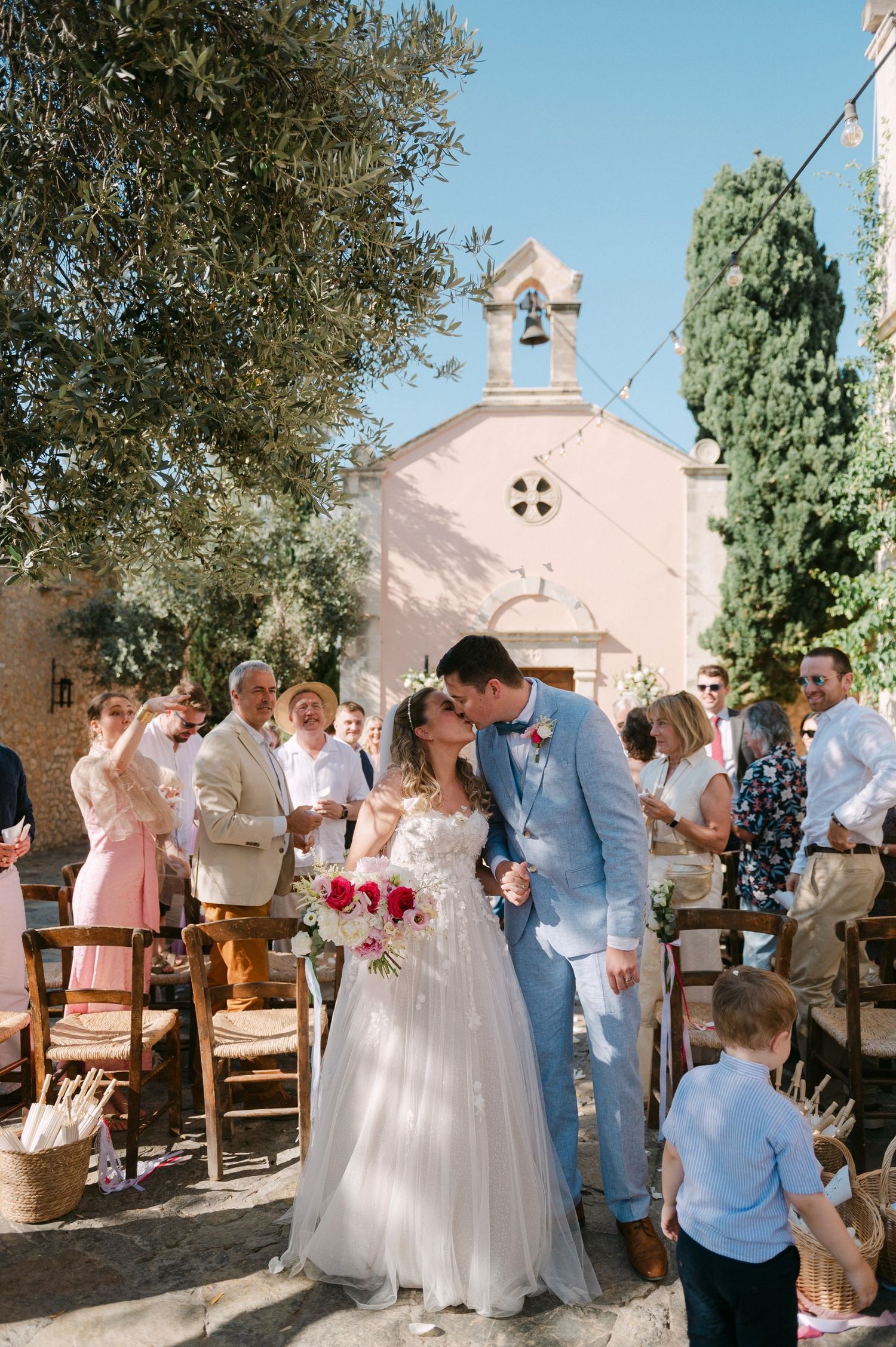 Wedding party gathered in front of a charming white adobe church on a sunny day during an outdoor celebration.