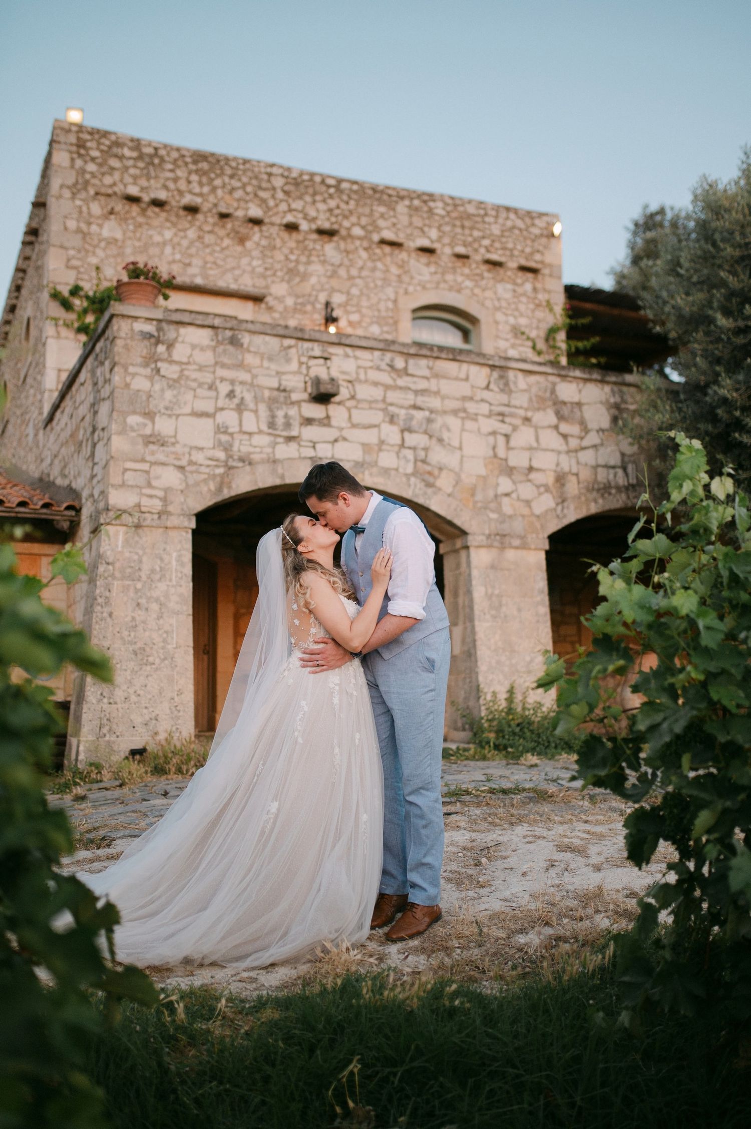 A couple shares romantic moments in wine country against stone walls and vineyard greenery at sunset.