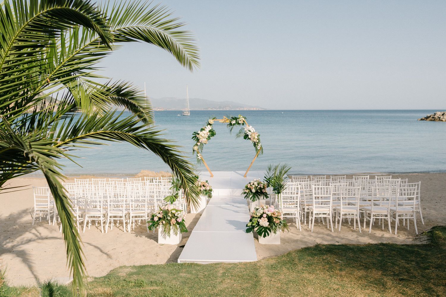 Beachfront wedding ceremony setup with white chairs and floral arch overlooking ocean.