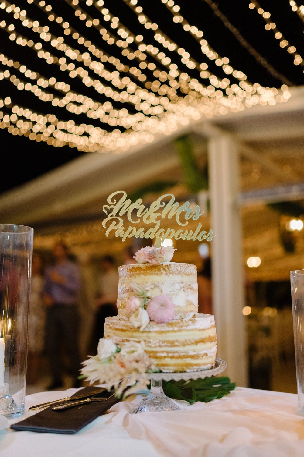 A wedding cake with floral decorations displayed under twinkling string lights at an outdoor reception.