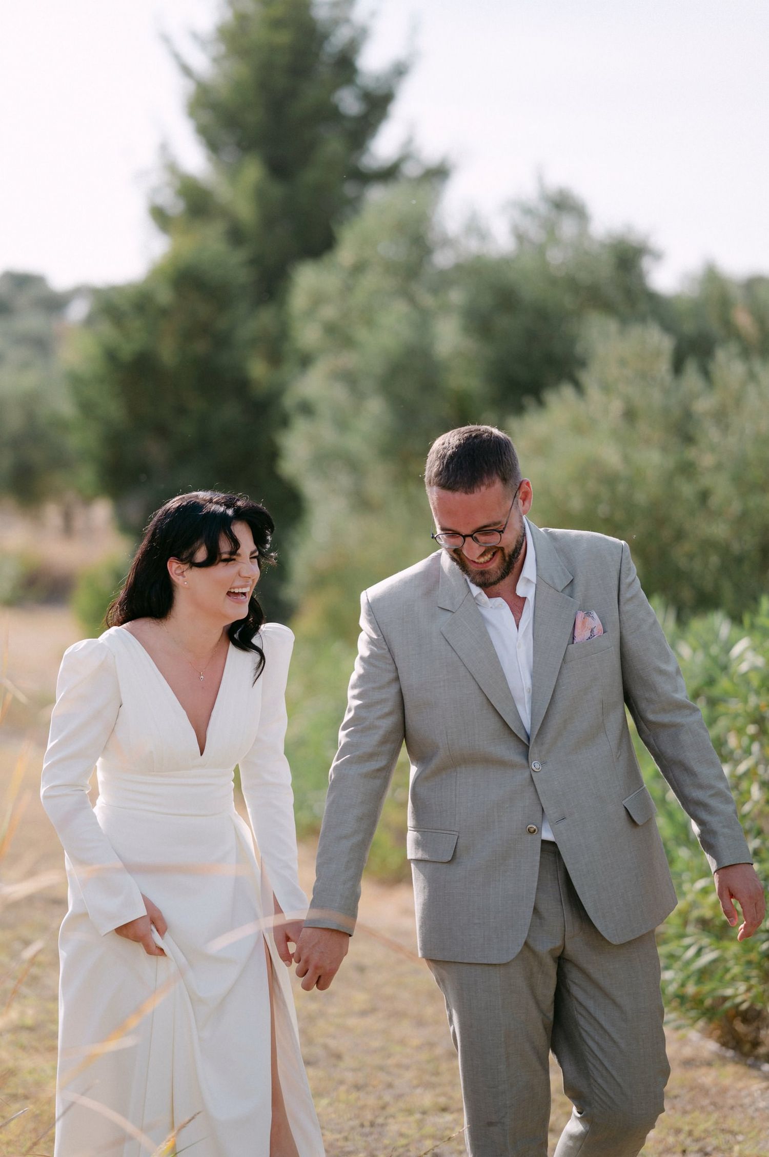 A couple in wedding attire walks hand in hand on a dirt path surrounded by greenery on a sunny day.