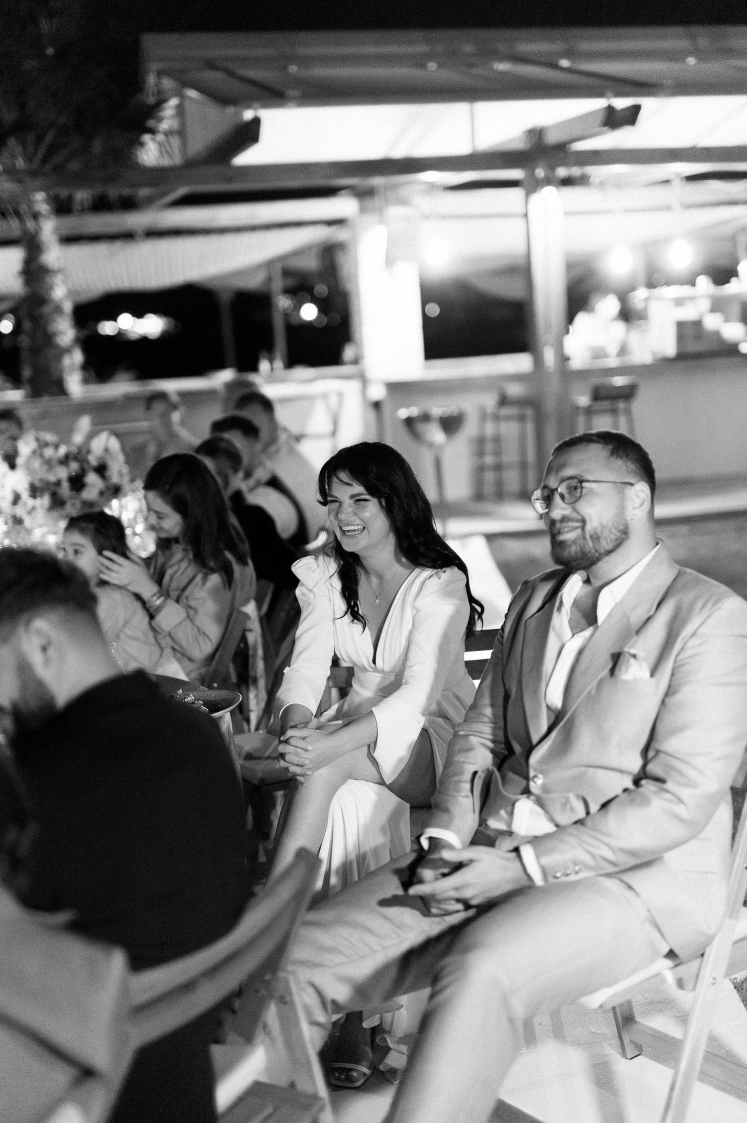 Wedding guests in formal attire seated at a reception raise their glasses in a celebratory toast.