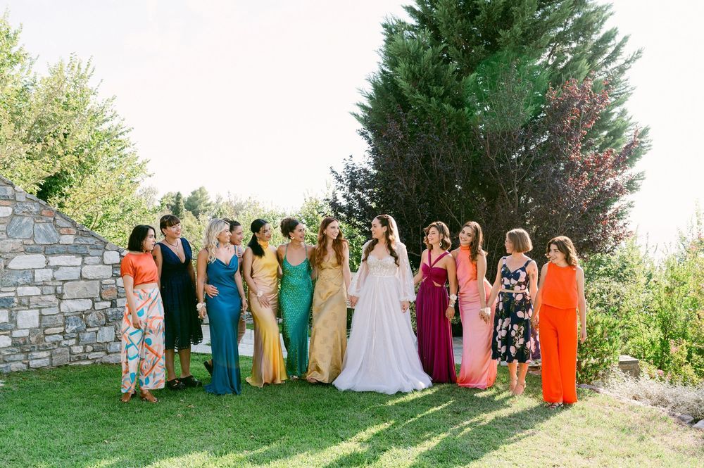 A group of people in colorful formal dresses and gowns stand together on a grassy lawn near stone wall and evergreen trees.