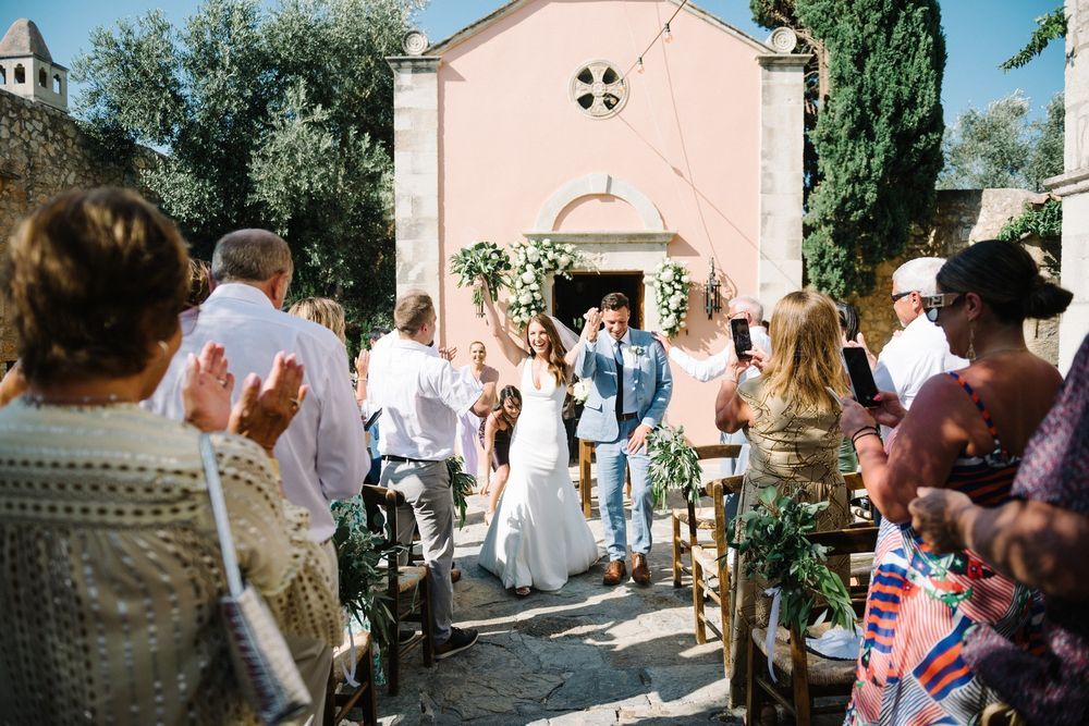 A pink church with a bell tower stands surrounded by greenery as people gather outside for a special occasion in Agreco Farms, Crete.