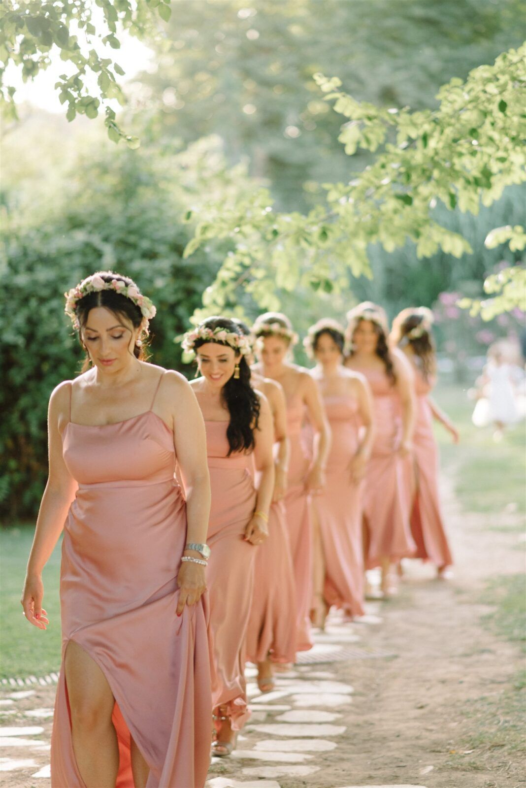 Bridesmaids in matching pink dresses walking in a line along a sunlit garden path at Parga, Greece
