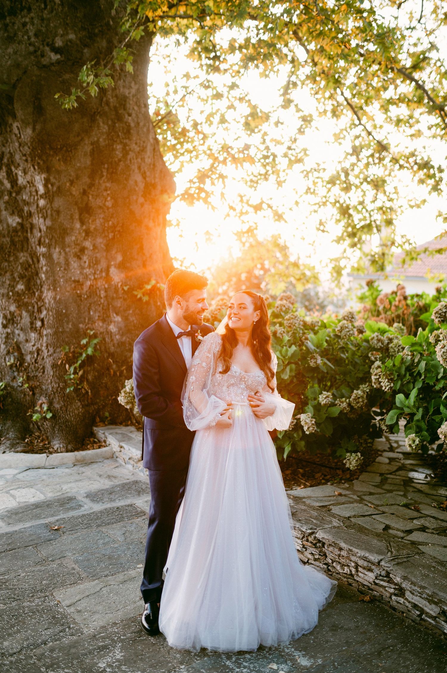 Romantic sunset wedding photo against stone wall with sunlight streaming through trees in golden hour lighting.