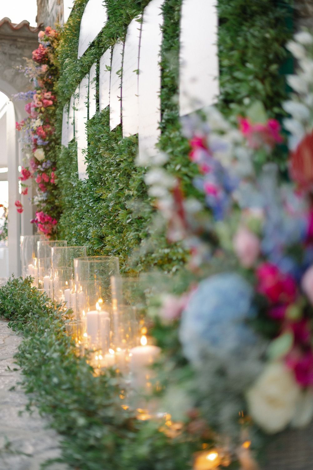 Candlelit pathway decorated with greenery and vibrant flowers along white garden fence.