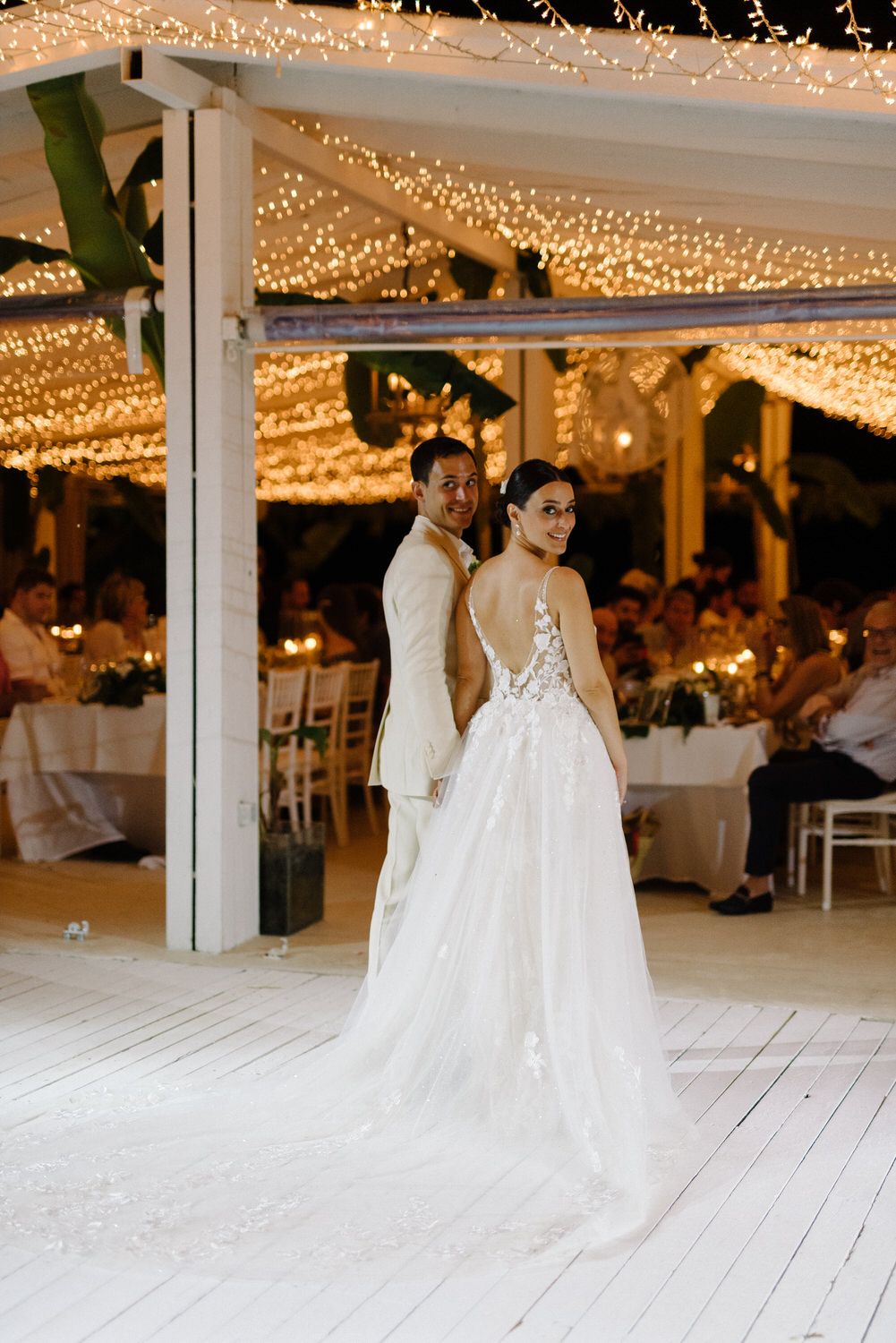 Elegant wedding couple standing on a white deck beneath a stunning canopy of golden fairy lights.