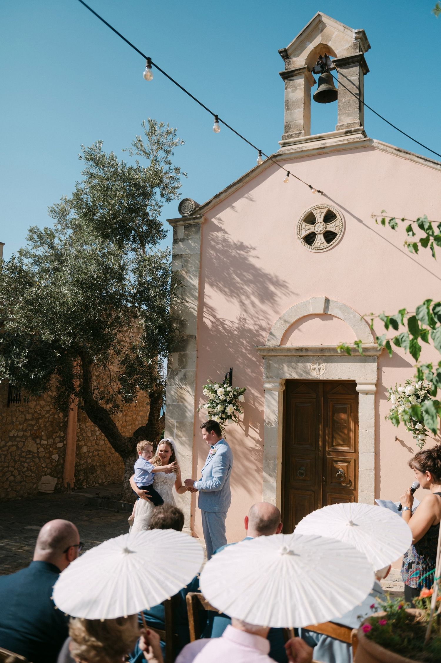 A pink Mediterranean church chapel with a bell tower and guests with white parasols gathered outside for a summer ceremony.