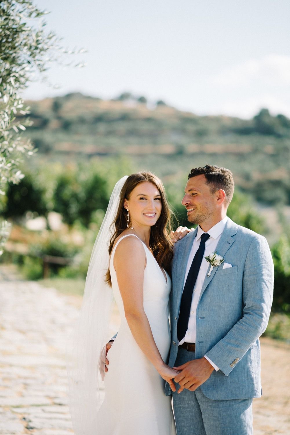 A happy wedding couple stands together outdoors in Tuscany, with the bride in white and groom in light blue suit.