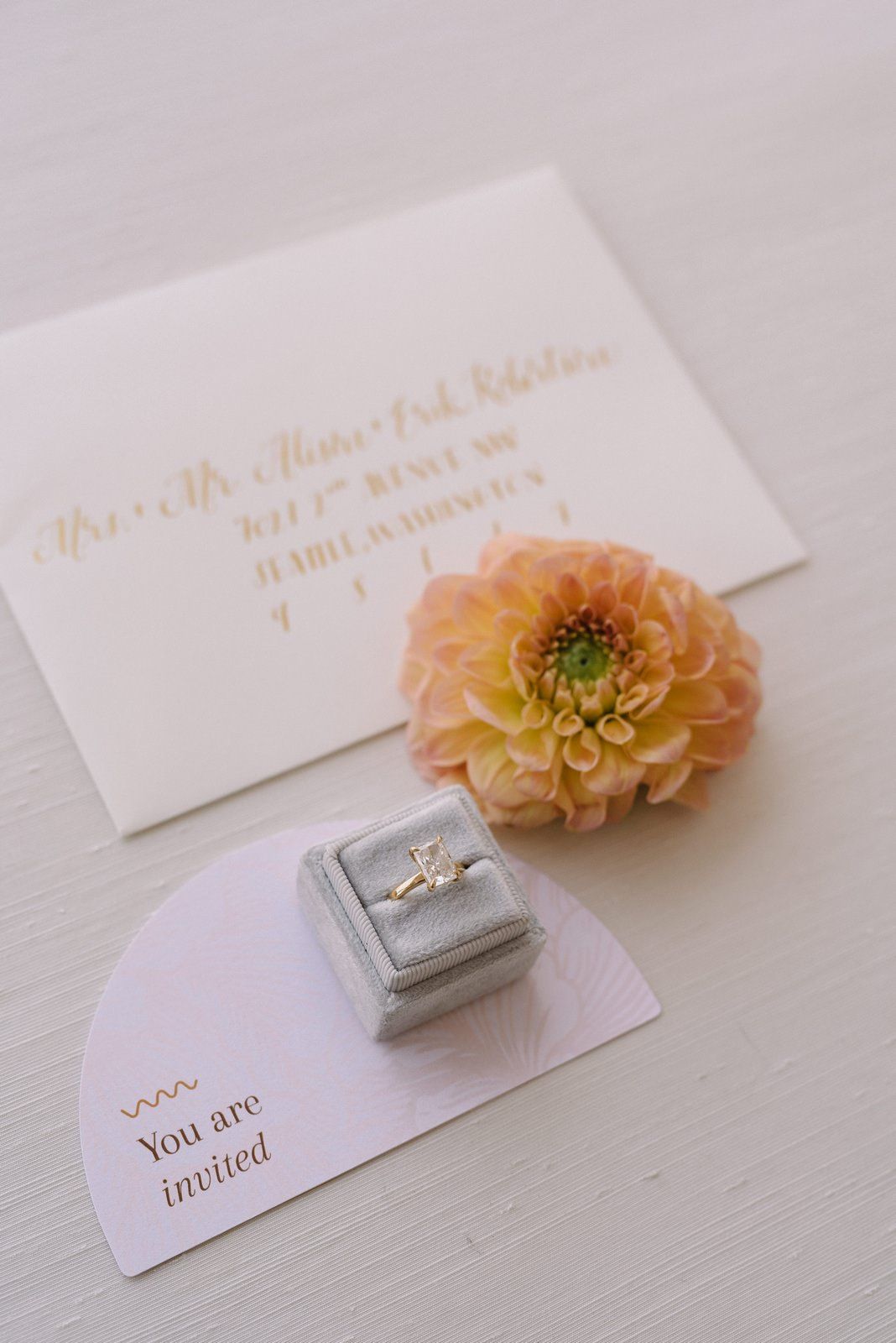 Wedding invitation with a peach dahlia flower and elegant diamond ring in grey velvet box.