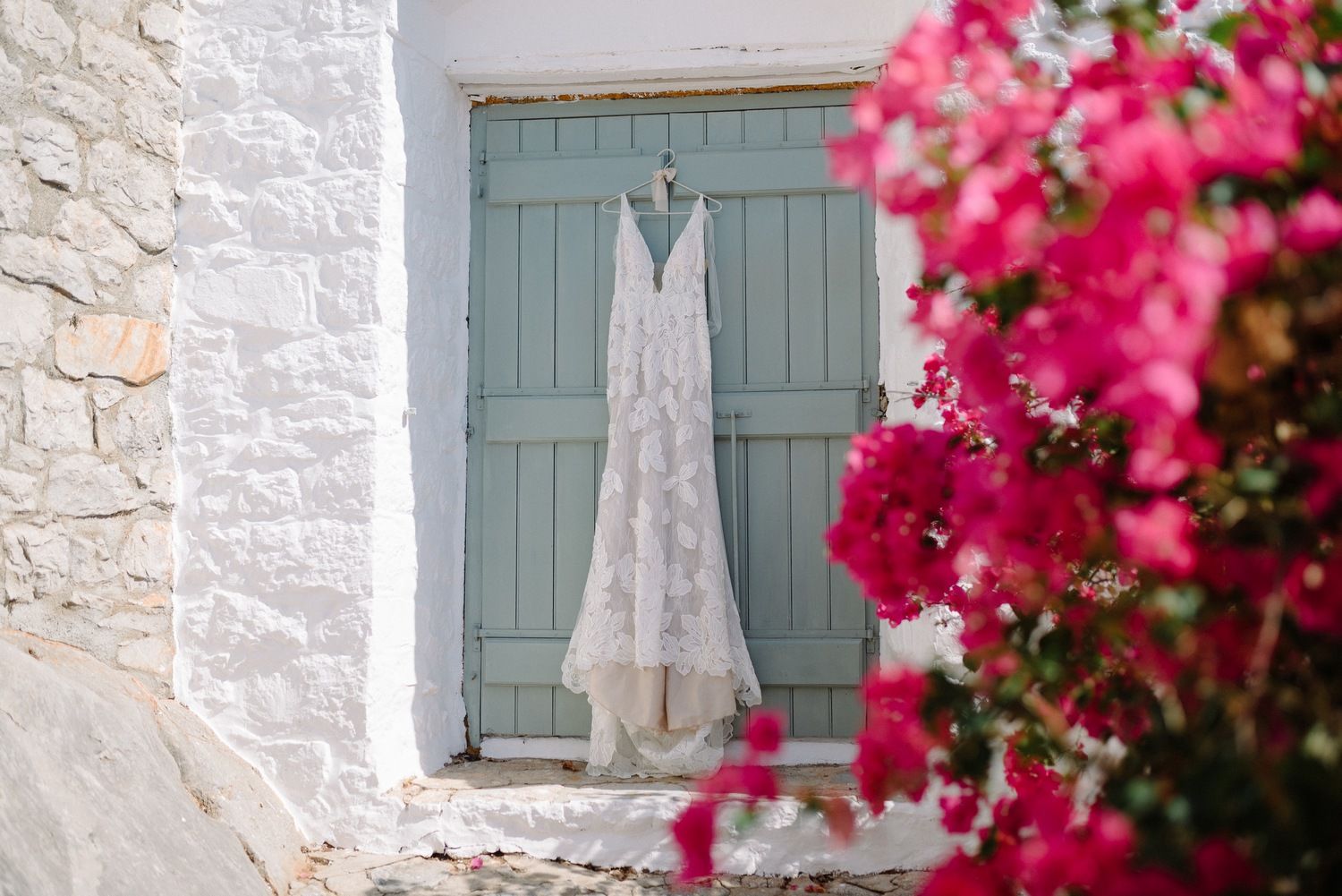 A white wedding dress hangs on a pale blue door against a whitewashed wall with pink bougainvillea flowers.